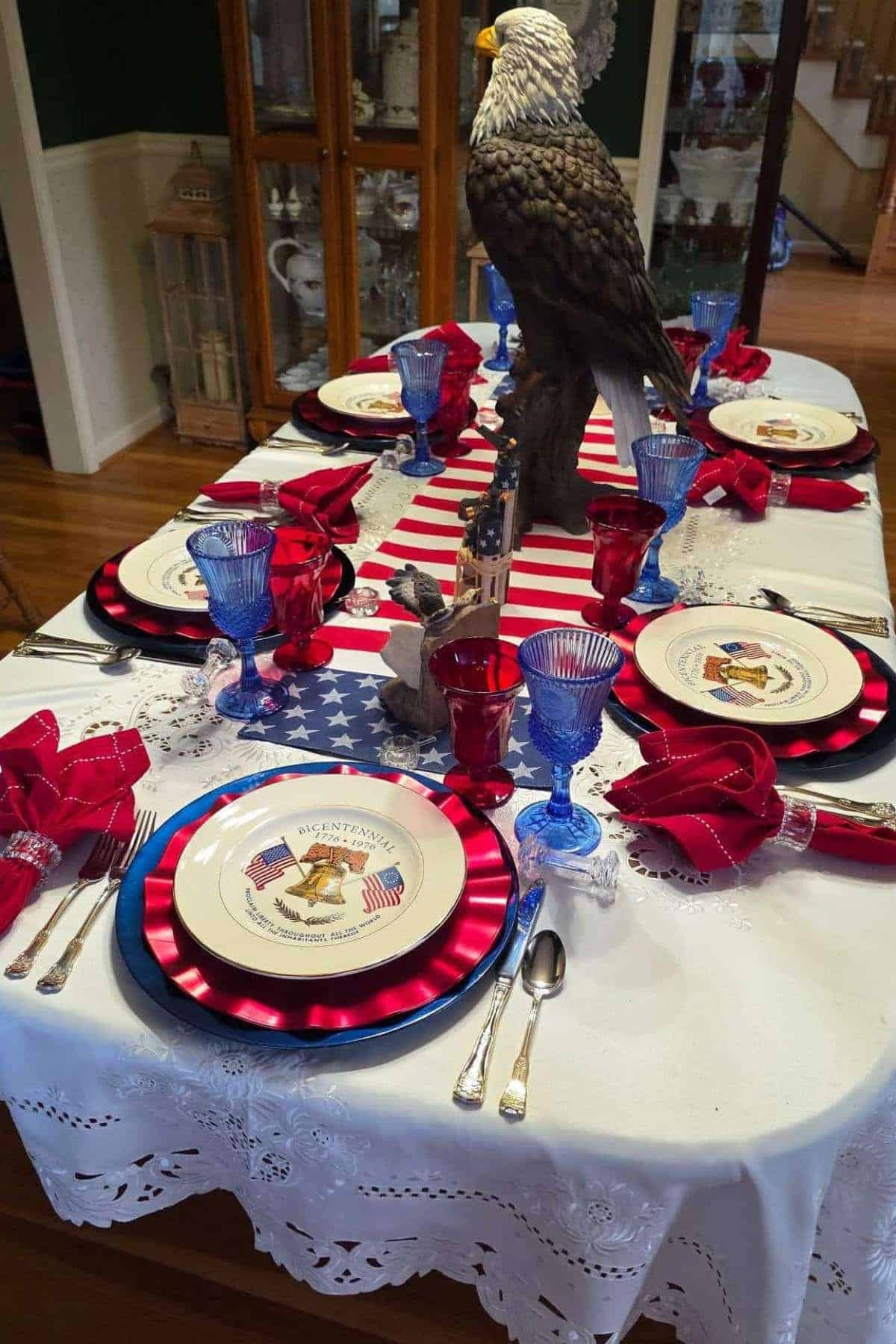 A dining table set with patriotic-themed plates, red and blue glasses, red napkins, and a large bald eagle centerpiece on a white lace tablecloth.