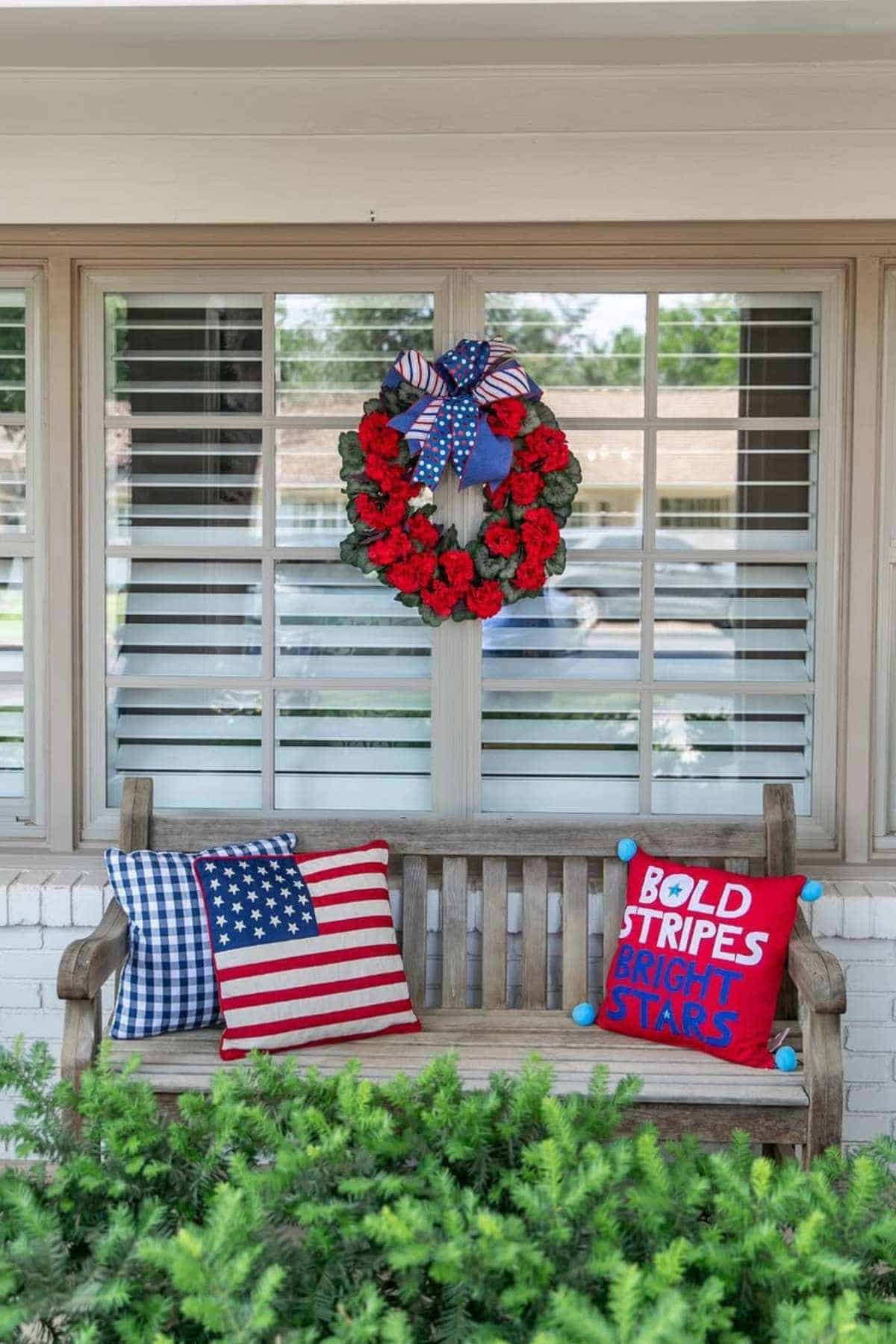 A wooden bench in front of a window displays patriotic pillows and a red flower wreath with a patriotic bow.