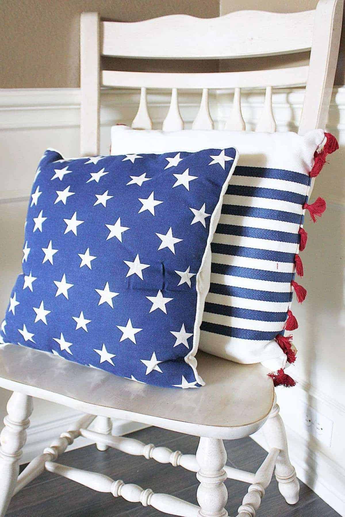 Two decorative pillows, one with white stars on a blue background and one with navy stripes and red tassels, are placed on a wooden chair.