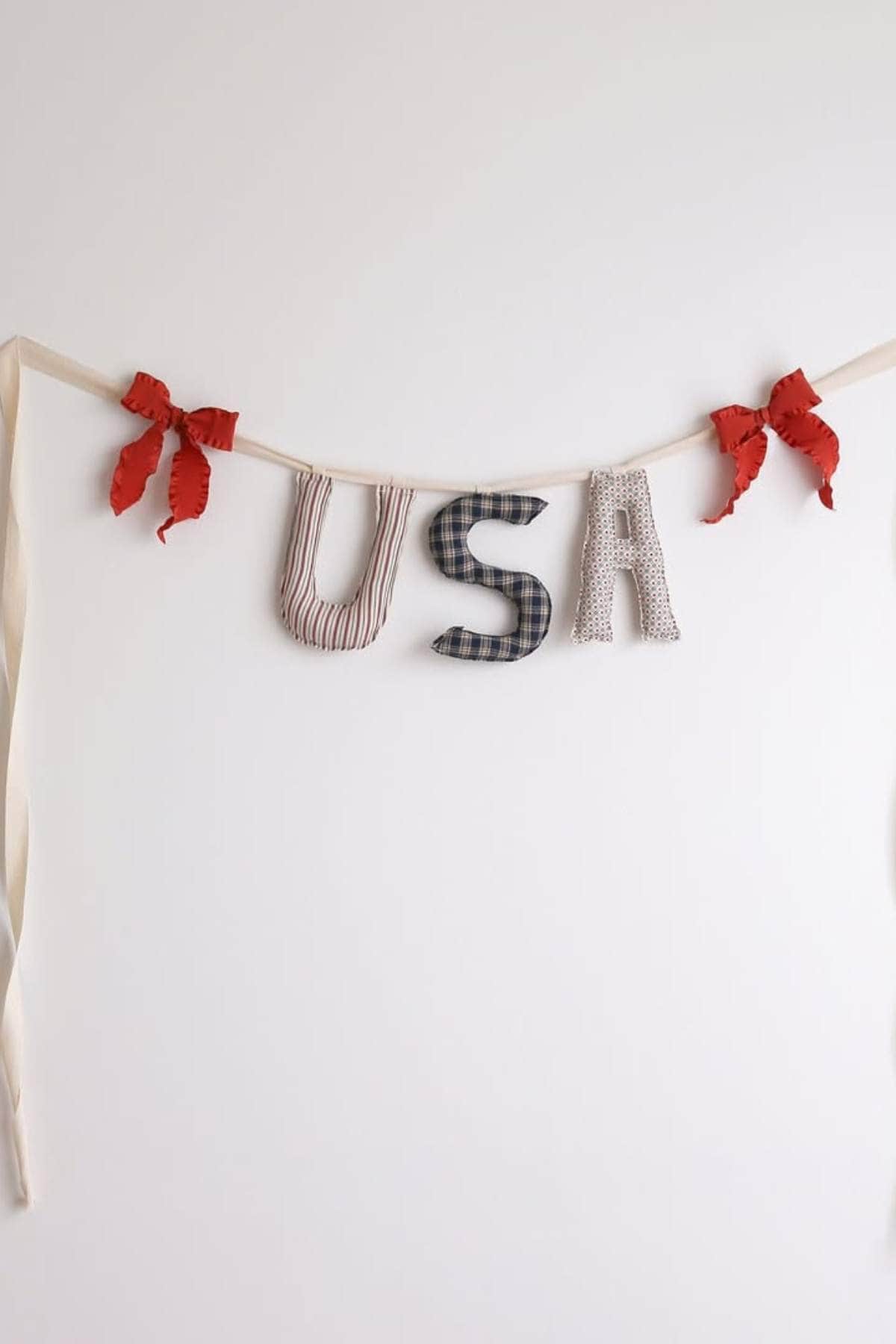 Fabric letters spelling "USA" hang from a cream-colored ribbon with two red bows on a plain white background.