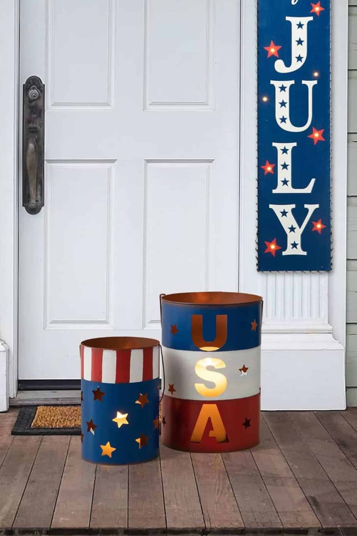 Two patriotic metal lanterns, one with stars and stripes, the other spelling "USA" with cutouts, sit on a porch in front of a white door.