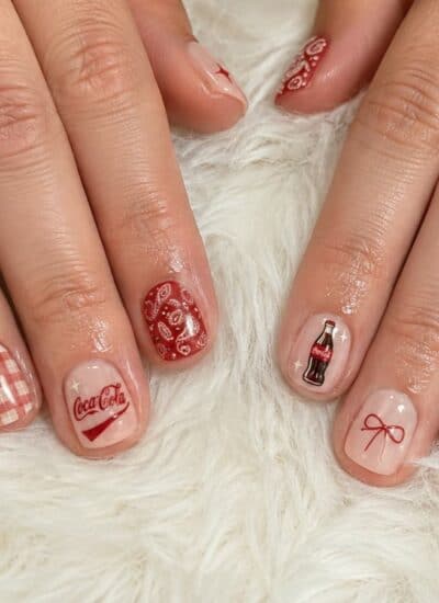 Hands with nail art featuring red and white gingham patterns, cherries, a Coca-Cola logo, a soda bottle, a bow, and a bandana print design on a white furry surface.