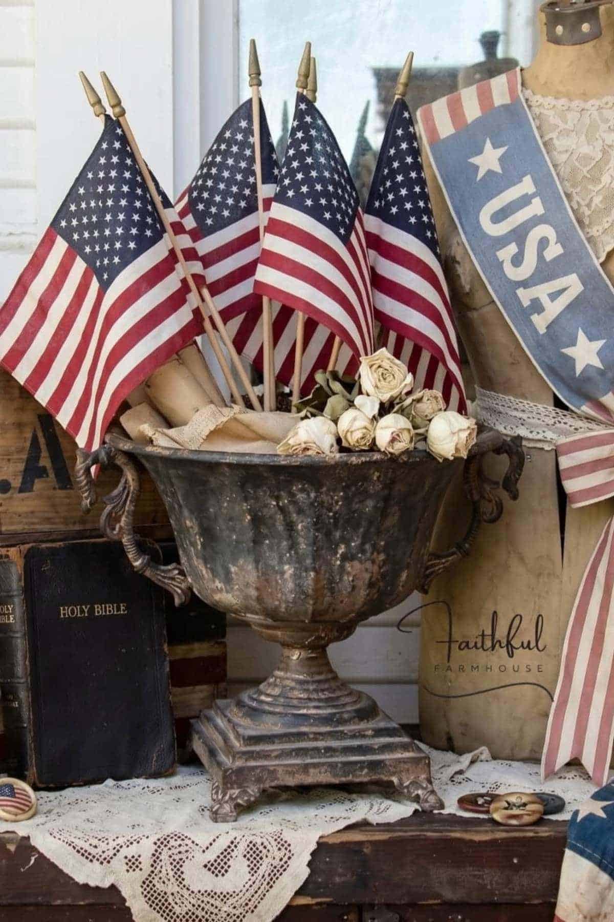 A tarnished metal urn holds several small American flags and white roses, surrounded by patriotic decorations, an old Holy Bible, and lace on a wooden table.
