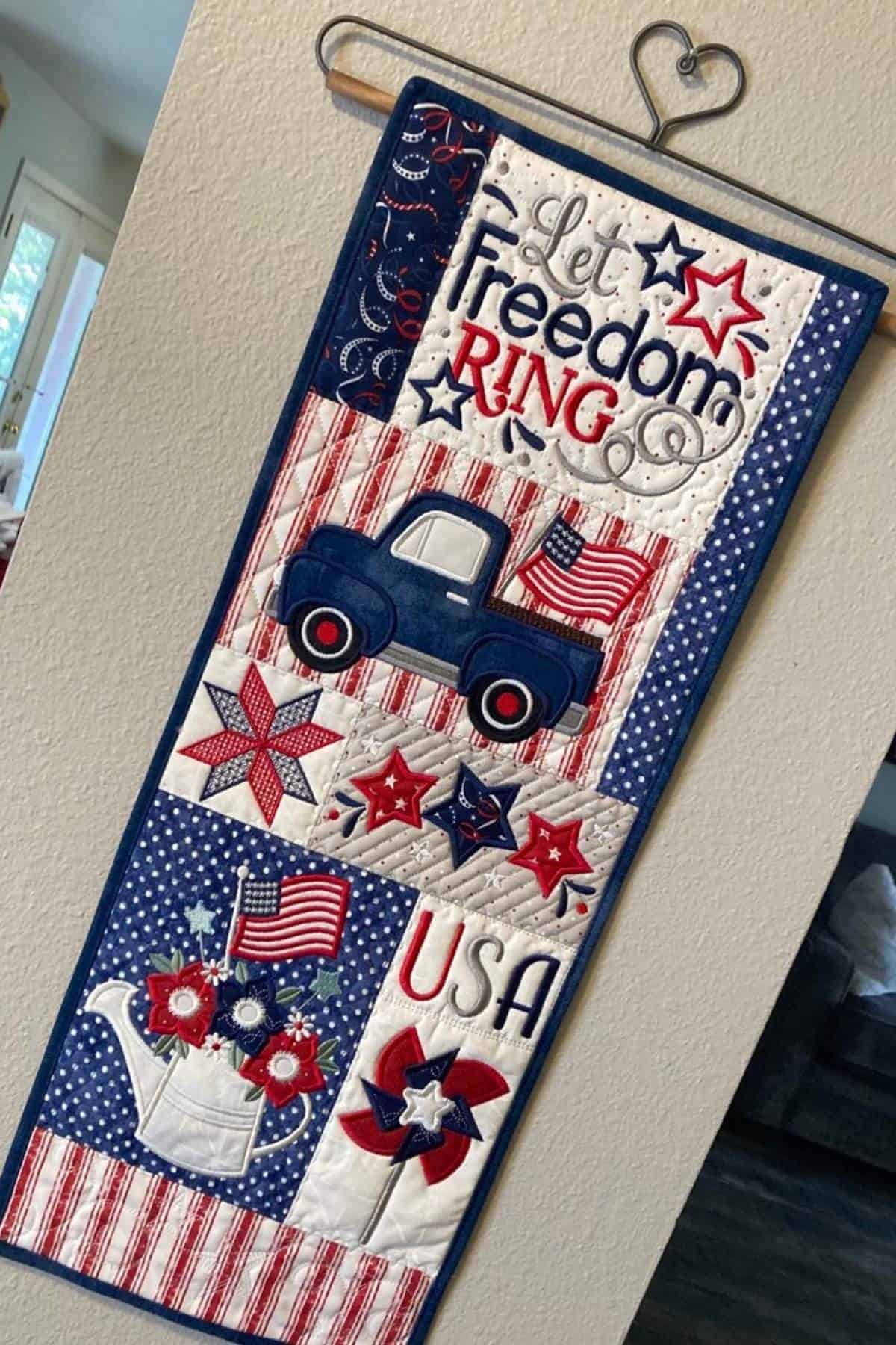 A decorative quilted wall hanging with patriotic motifs, including a truck, flowers, American flags, stars, and the words "Let Freedom Ring" and "USA.