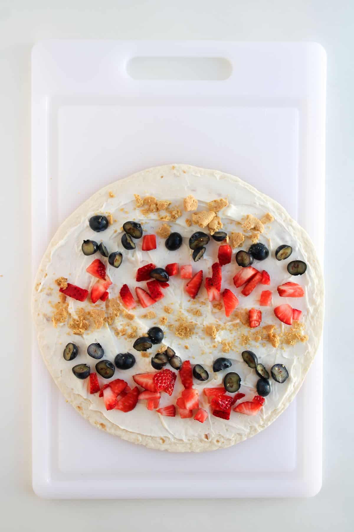 A tortilla topped with a layer of cream, blueberries, chopped strawberries, and crumbled graham crackers on a white cutting board.