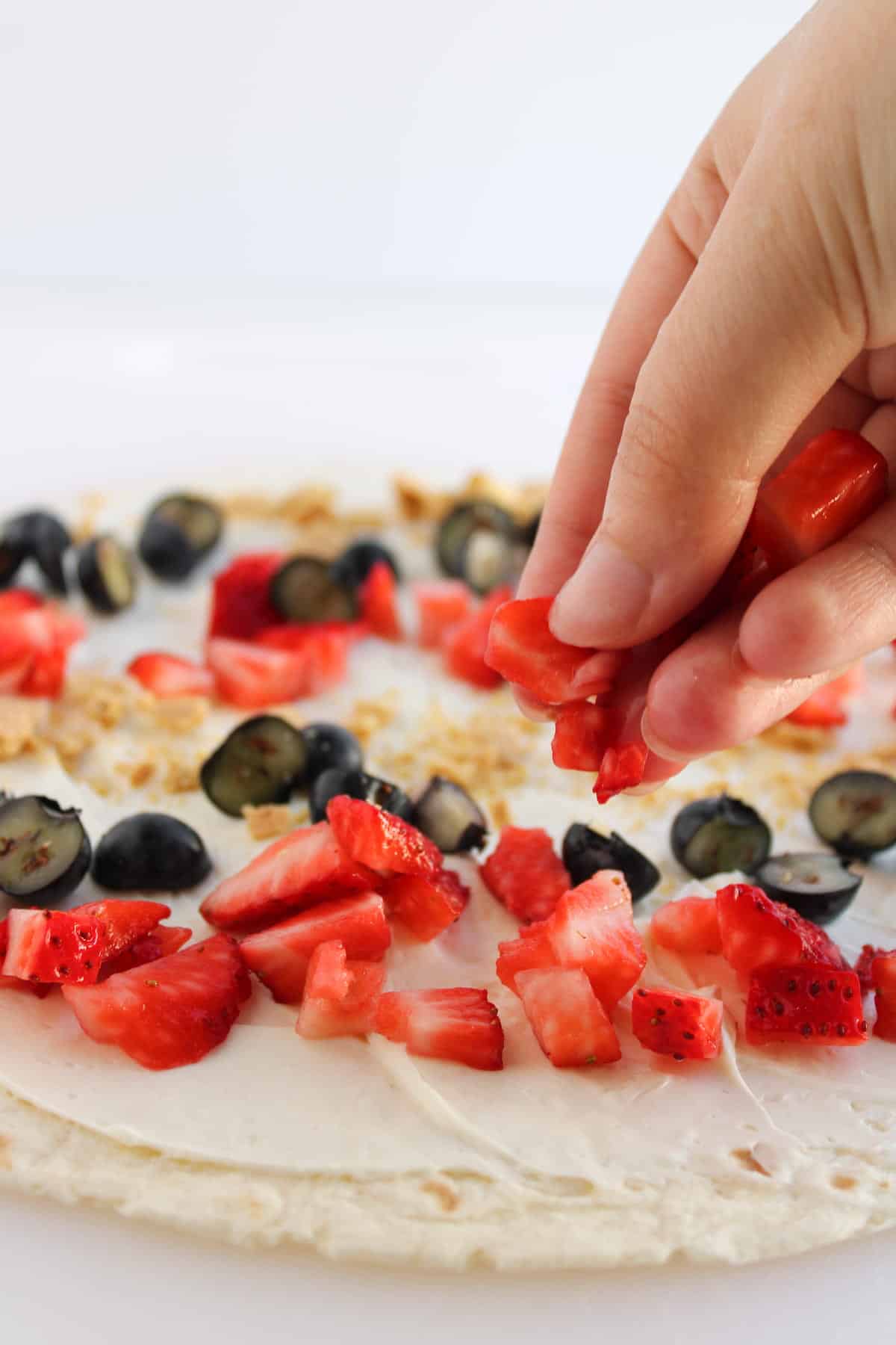 A hand adds chopped strawberries to a tortilla spread with cream cheese, also topped with blueberries and crushed graham crackers.