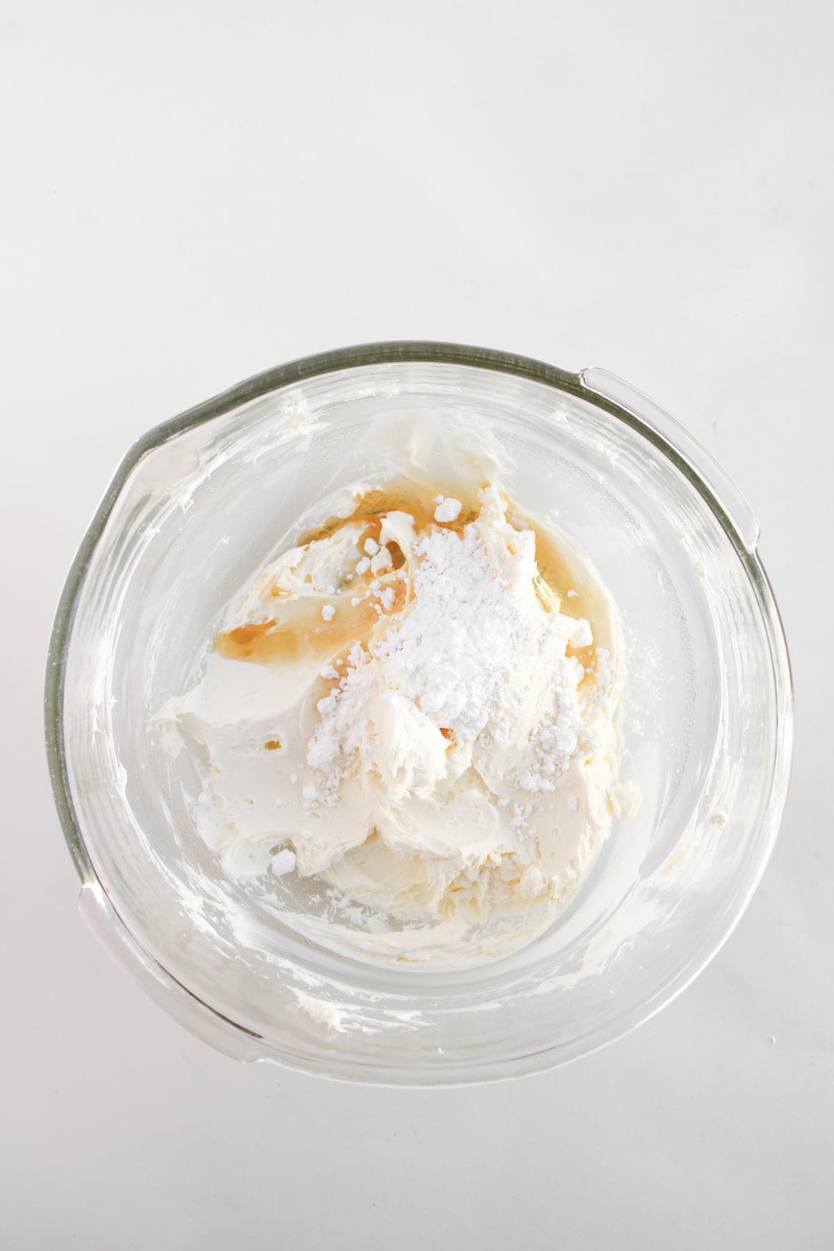 A glass mixing bowl containing cream cheese, powdered sugar, and vanilla extract on a white surface.