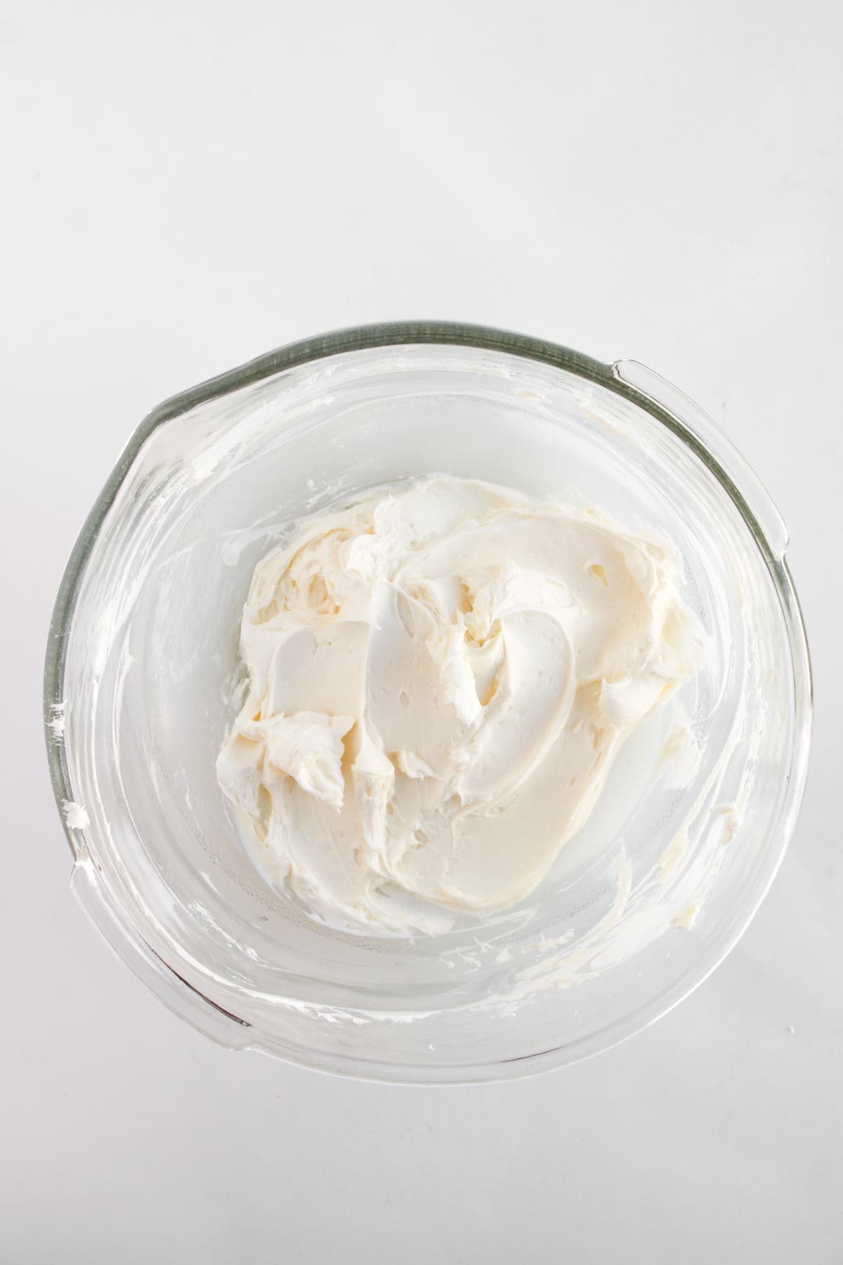 A glass mixing bowl filled with smooth, white whipped frosting on a white background.