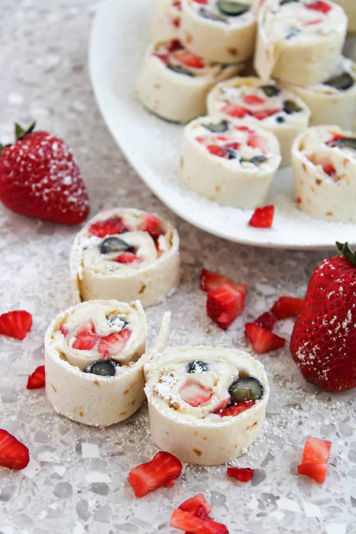 Sliced tortilla rolls filled with cream cheese, strawberries, and blueberries are arranged on a plate and countertop, with fresh strawberries and diced fruit scattered around.