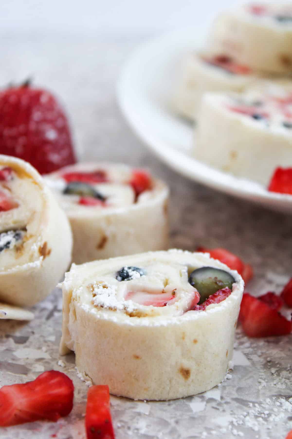 Close-up of fruit and cream cheese pinwheels made with tortilla, sliced and arranged on a surface with pieces of strawberry and a plate in the background.
