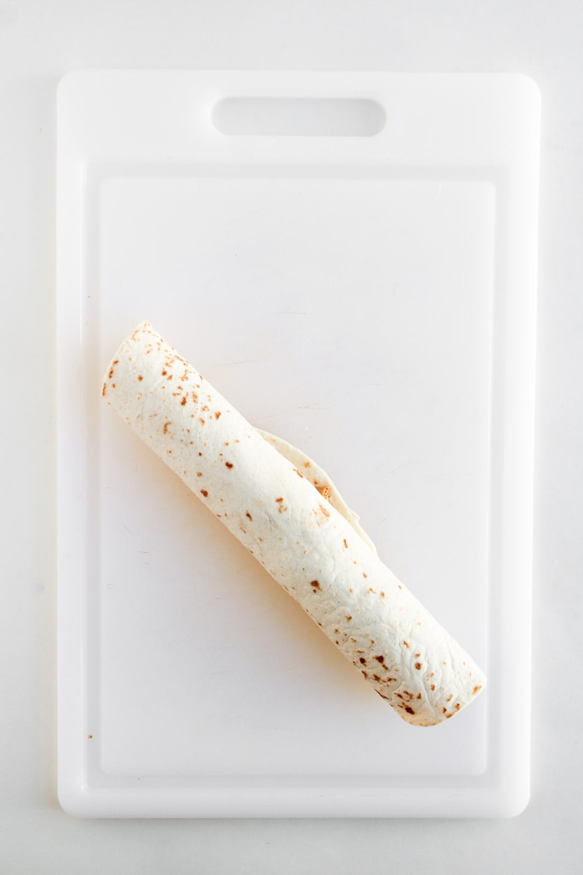 A rolled flour tortilla filled with ingredients sits centered on a white plastic cutting board against a plain background.
