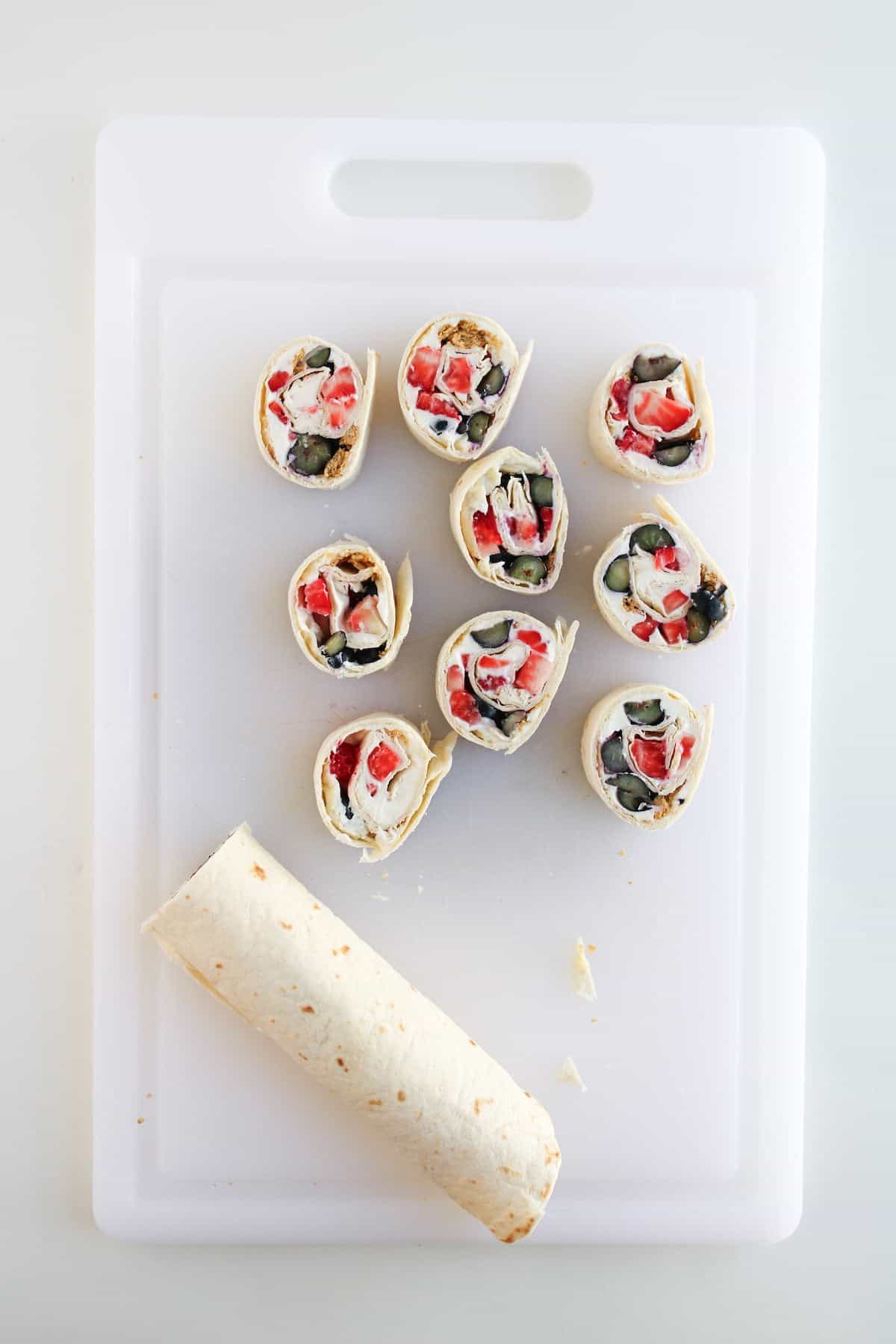 A sliced tortilla wrap with colorful vegetable filling is arranged on a white cutting board, with one unsliced wrap at the bottom.