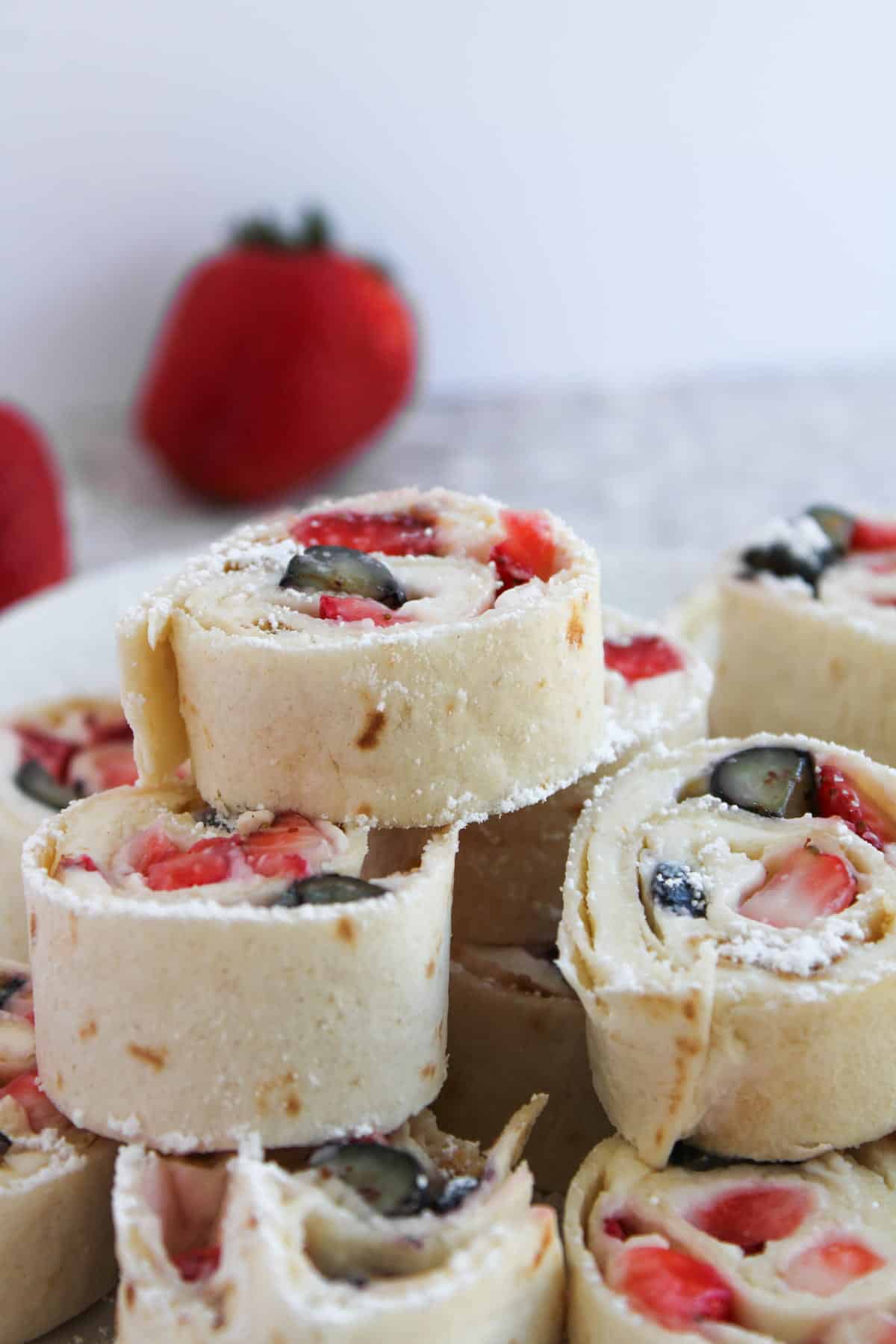 Close-up of tortilla roll-ups filled with cream, sliced strawberries, and blueberries, stacked on a plate with fresh strawberries in the background.
