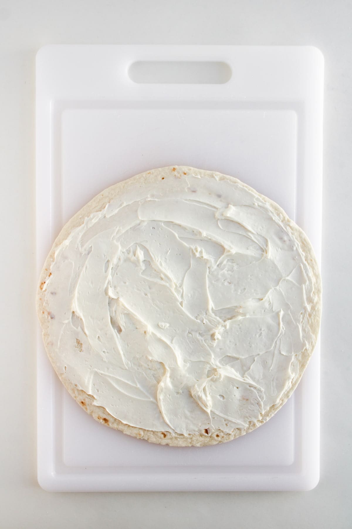 A large round tortilla spread with a layer of cream cheese sits on a white cutting board.