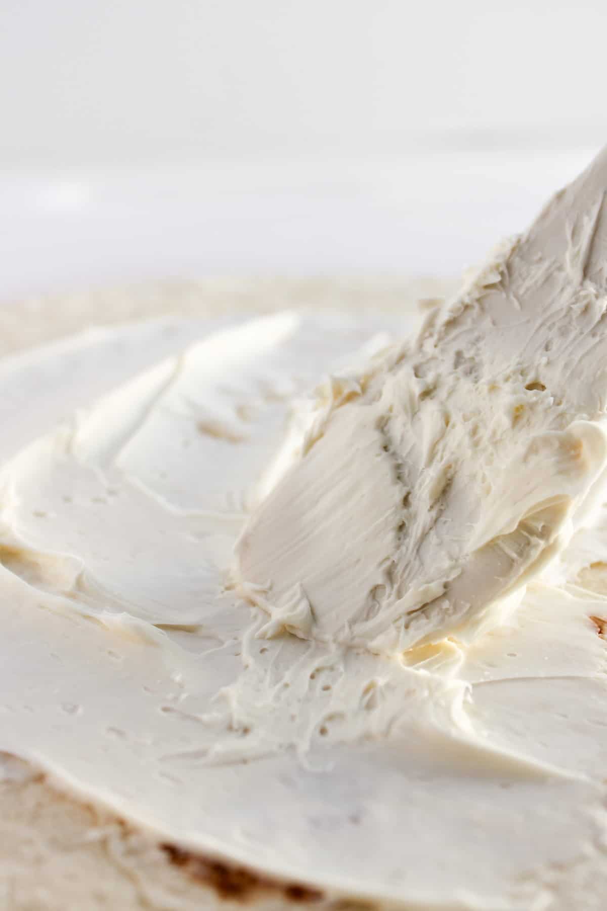 A close-up of a spatula spreading cream cheese or a similar white spread onto a tortilla.