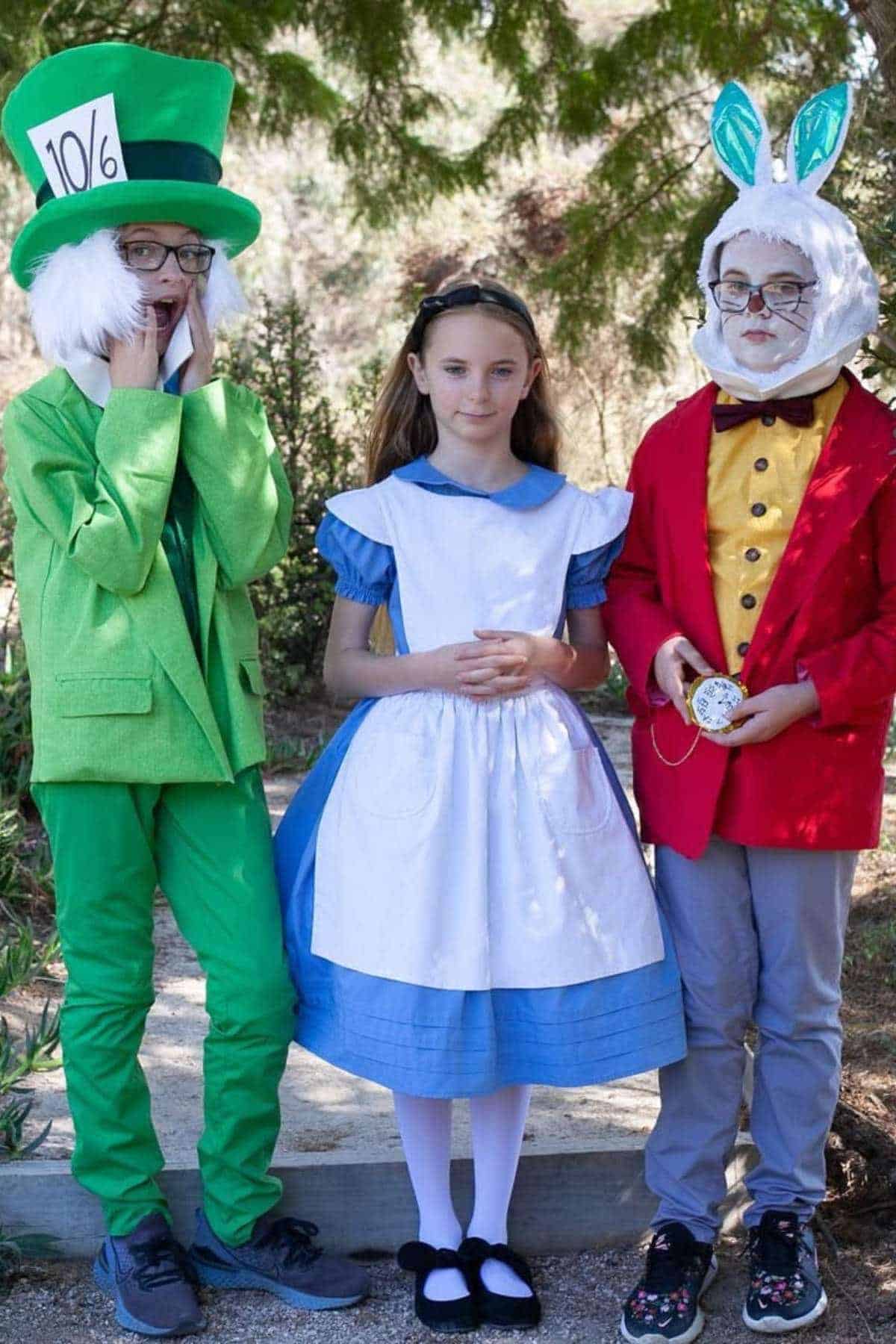 Three children dressed in costumes: one as the Mad Hatter, one as Alice, and one as the White Rabbit from Alice in Wonderland, standing outdoors.