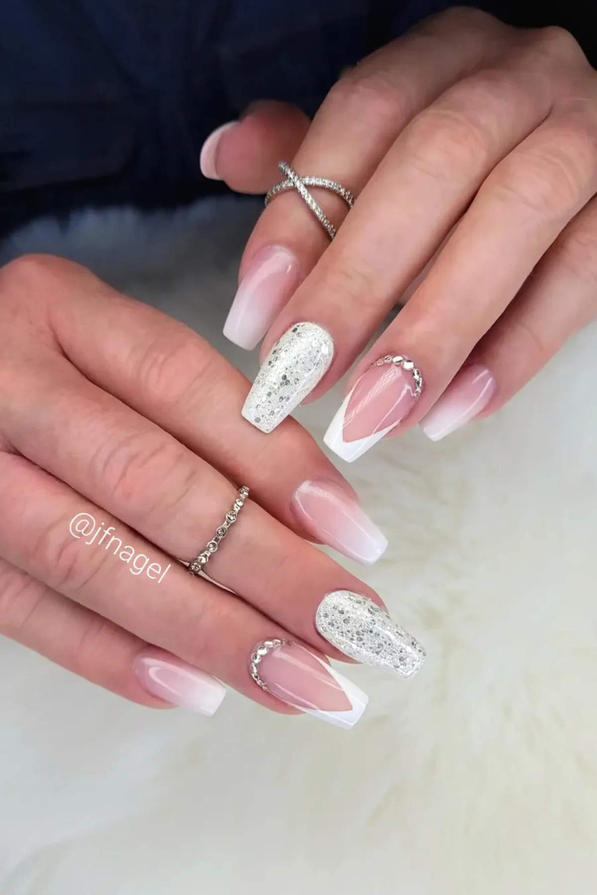 Hands with long coffin French manicure nails, featuring glitter accent nails and silver rhinestone decorations, resting on a white surface.
