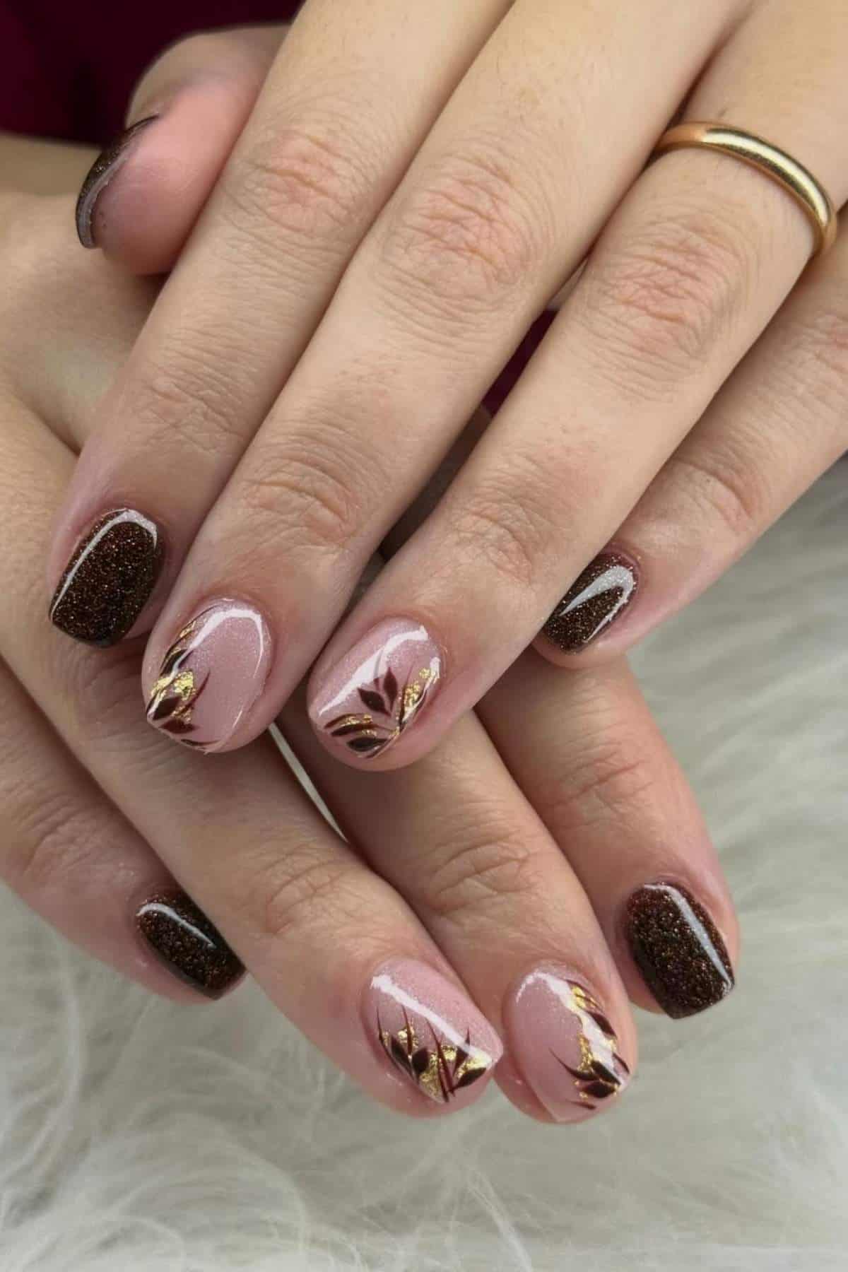 Close-up of hands with manicured nails featuring a mix of pink polish with gold leaf accents and dark brown glitter polish.