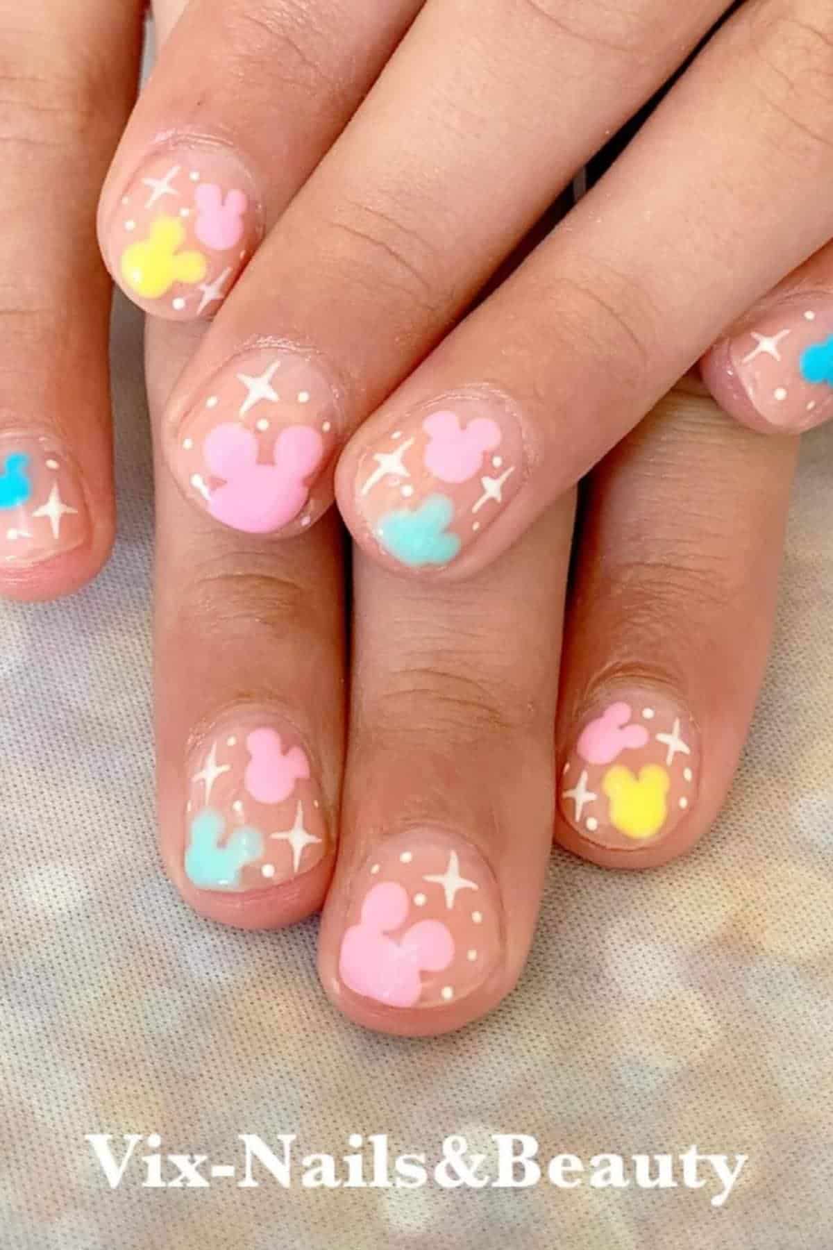 A hand with short nails painted with pastel Mickey Mouse head shapes and white star accents on a nude base. The text "Vix-Nails&Beauty" is overlaid at the bottom.