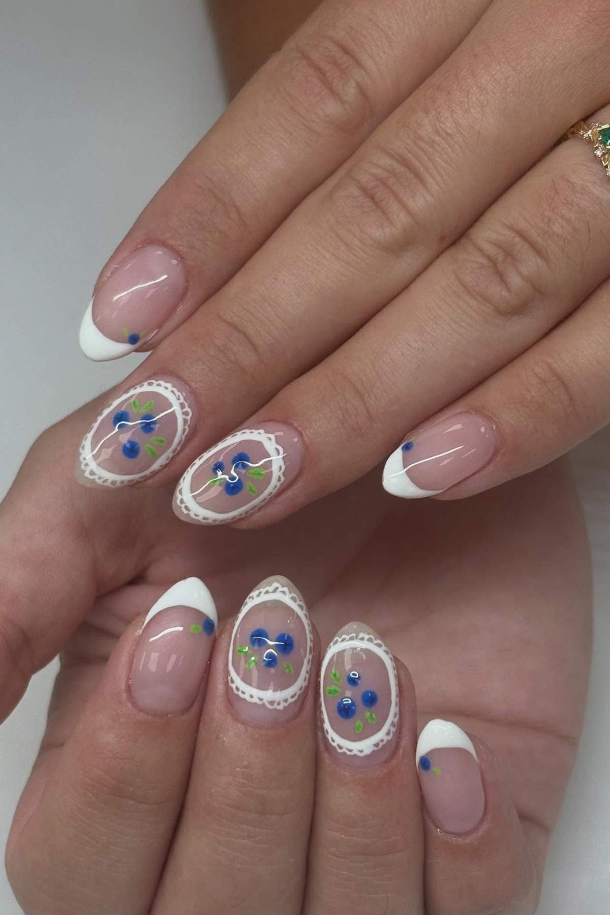 Hands with almond-shaped nails featuring French tips and white lace designs, blue flower nail art, and green leaf details on some nails.