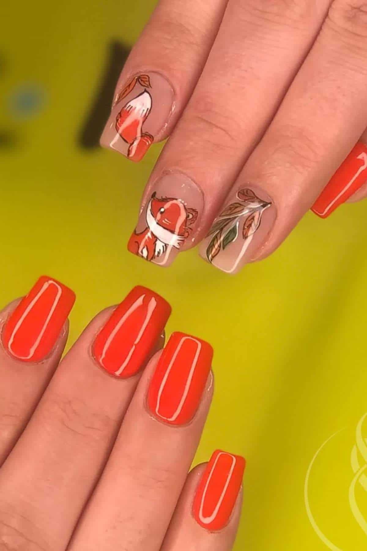 Hands with bright orange square-shaped nails; two accent nails feature a fox illustration and leaf designs on a nude base.