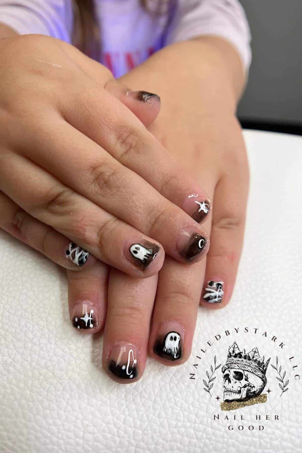 Child's hands with short nails painted with black, white, and brown Halloween-themed designs including ghosts, spider webs, and eyes.