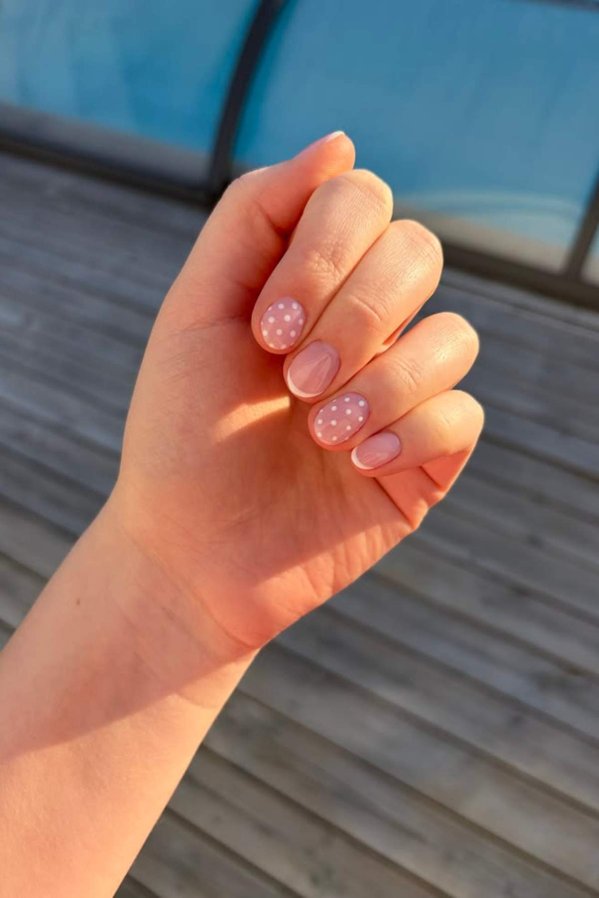 A hand with short, rounded nails painted light pink, with white polka dots on two fingernails, is held up against a wooden deck background.
