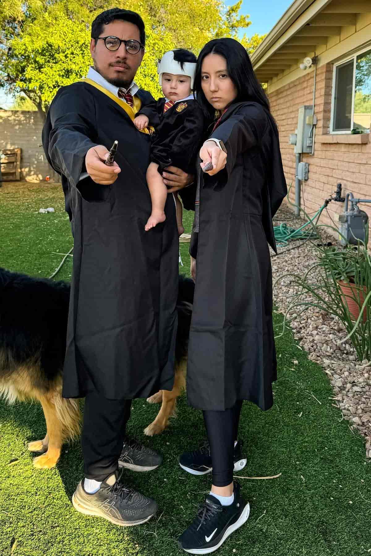 Three people in black robes and holding wands stand outside; two adults point wands forward while holding a baby. A dog stands beside them.