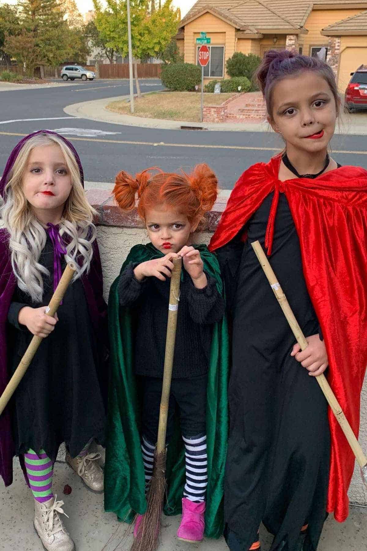 Three children dressed as witches in black outfits and colored capes stand outside holding brooms, with houses and a street in the background.