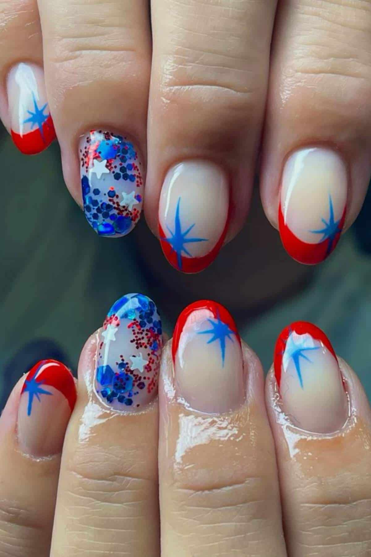 Hands with manicured nails featuring red tips, blue star designs, and accent nails with blue and red glitter on a nude base.