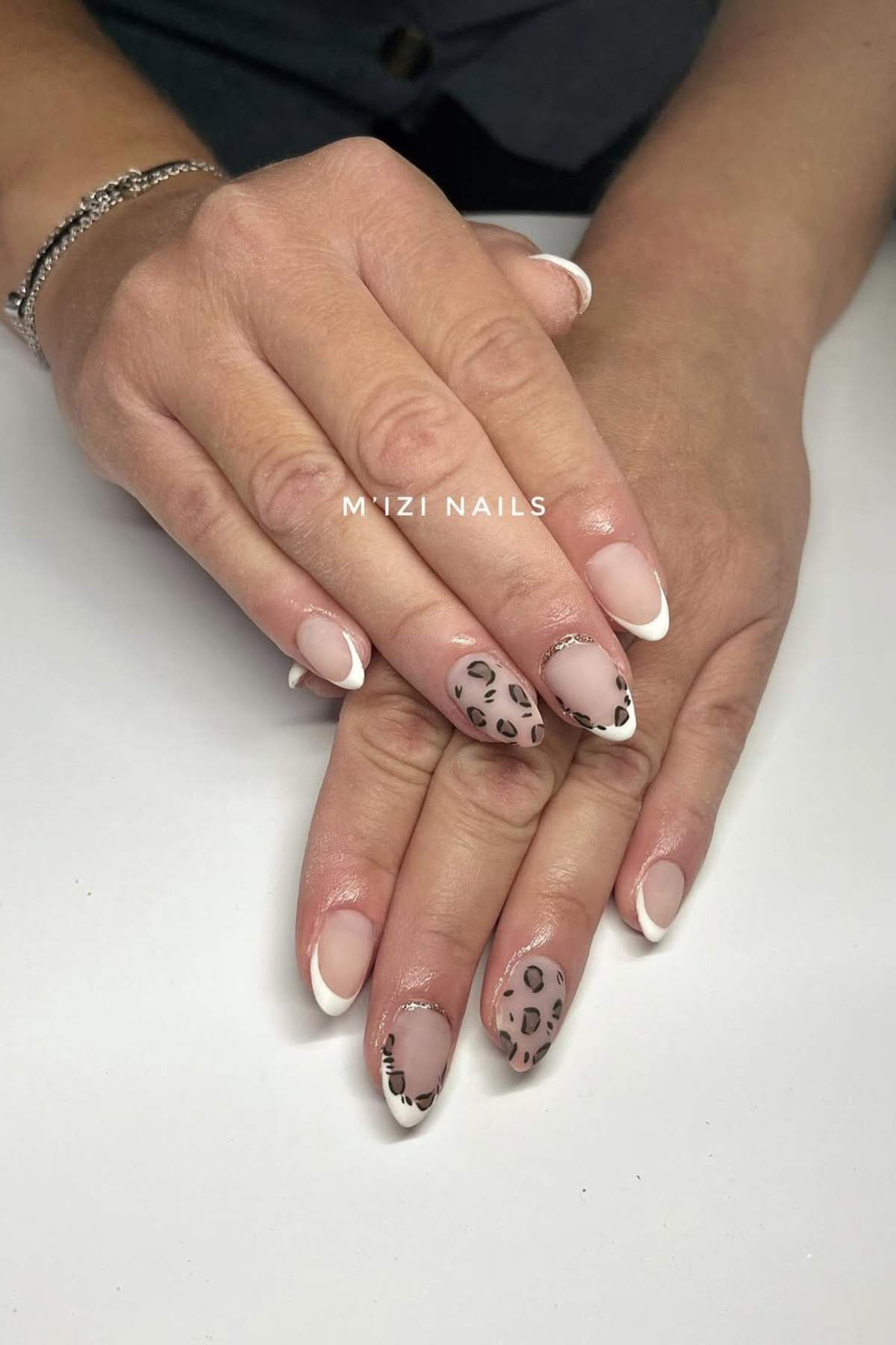 Hands with manicured nails featuring a mix of French tips and leopard print designs on a neutral base, resting on a white surface.