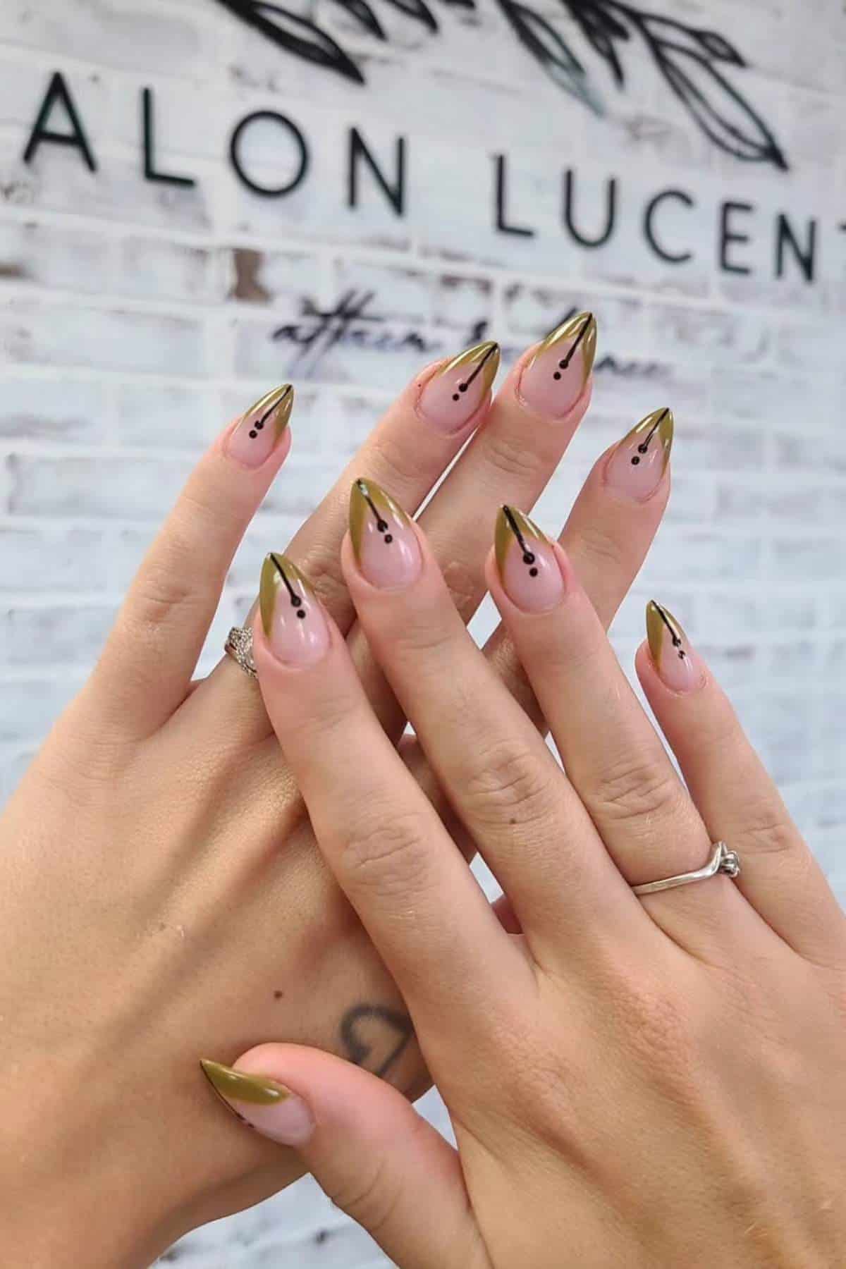 Hands with almond-shaped nails featuring a nude base, gold metallic tips, and black dot accents, shown against a blurred salon sign background.