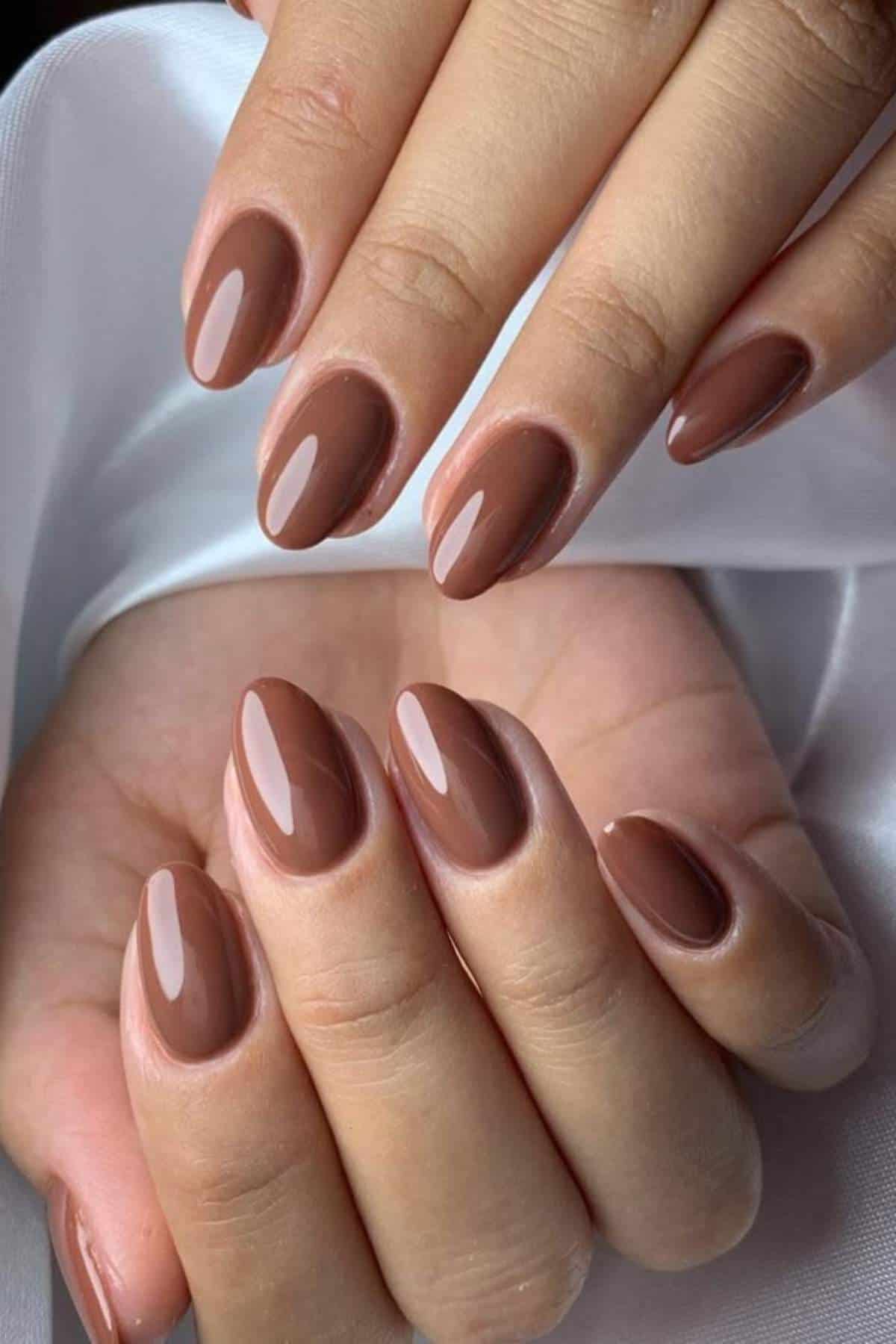 Close-up of hands showing neatly manicured nails painted in a glossy brown shade, with short, oval-shaped nails against a white fabric background.