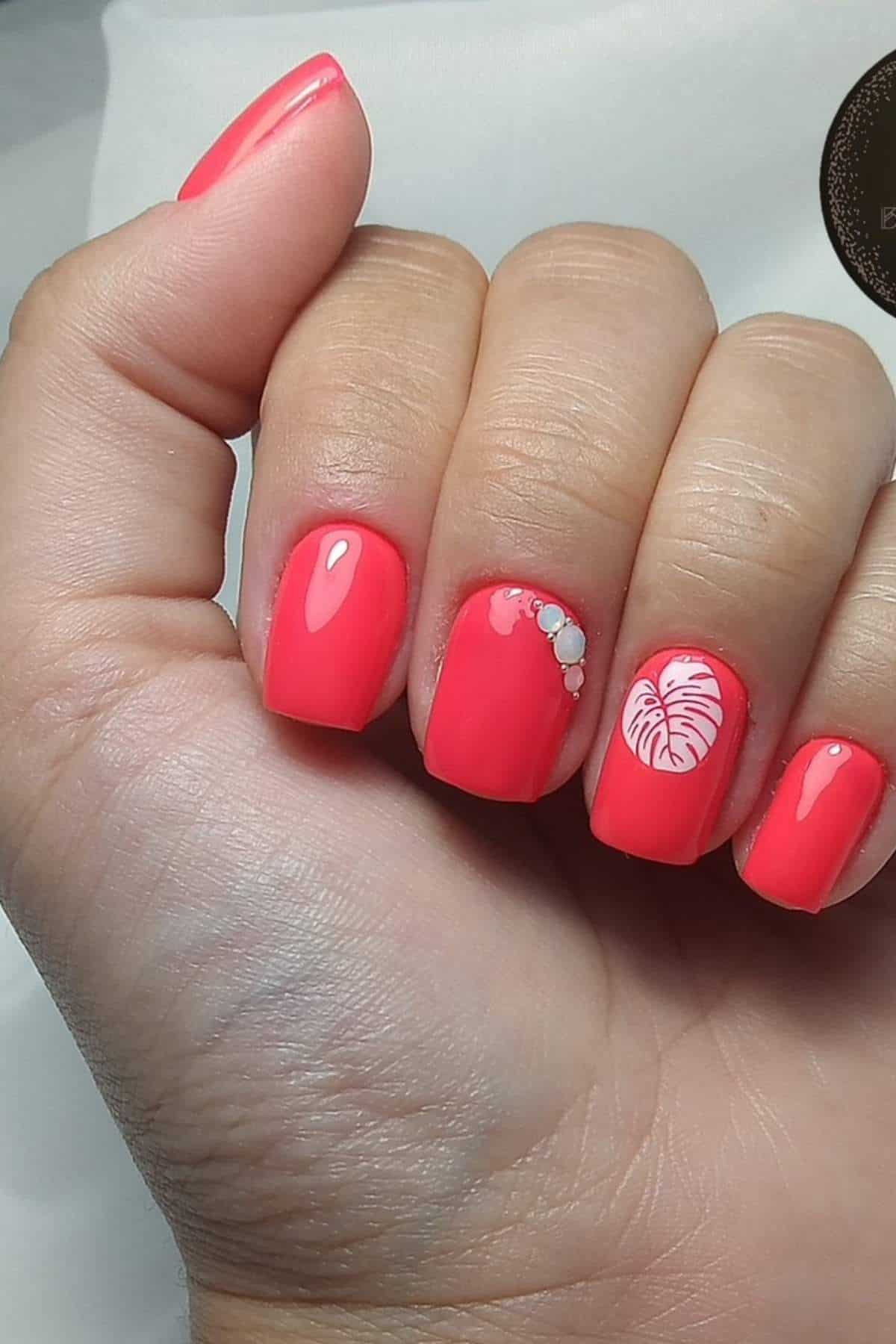 A hand with short coral-colored nails; one nail features a white leaf design and another has small pearl-like embellishments.