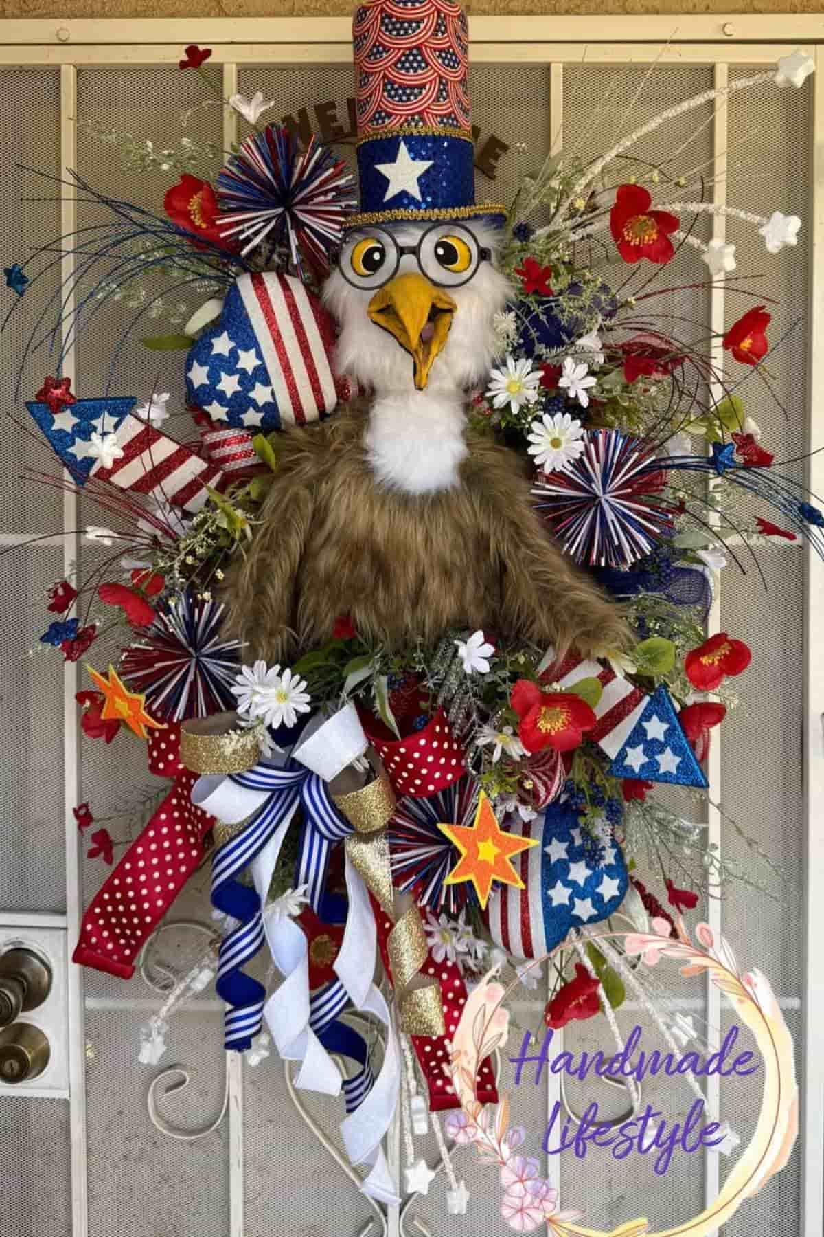 A patriotic door wreath featuring a plush eagle wearing a stars-and-stripes hat, surrounded by red, white, and blue ribbons, artificial flowers, and decorative accents.