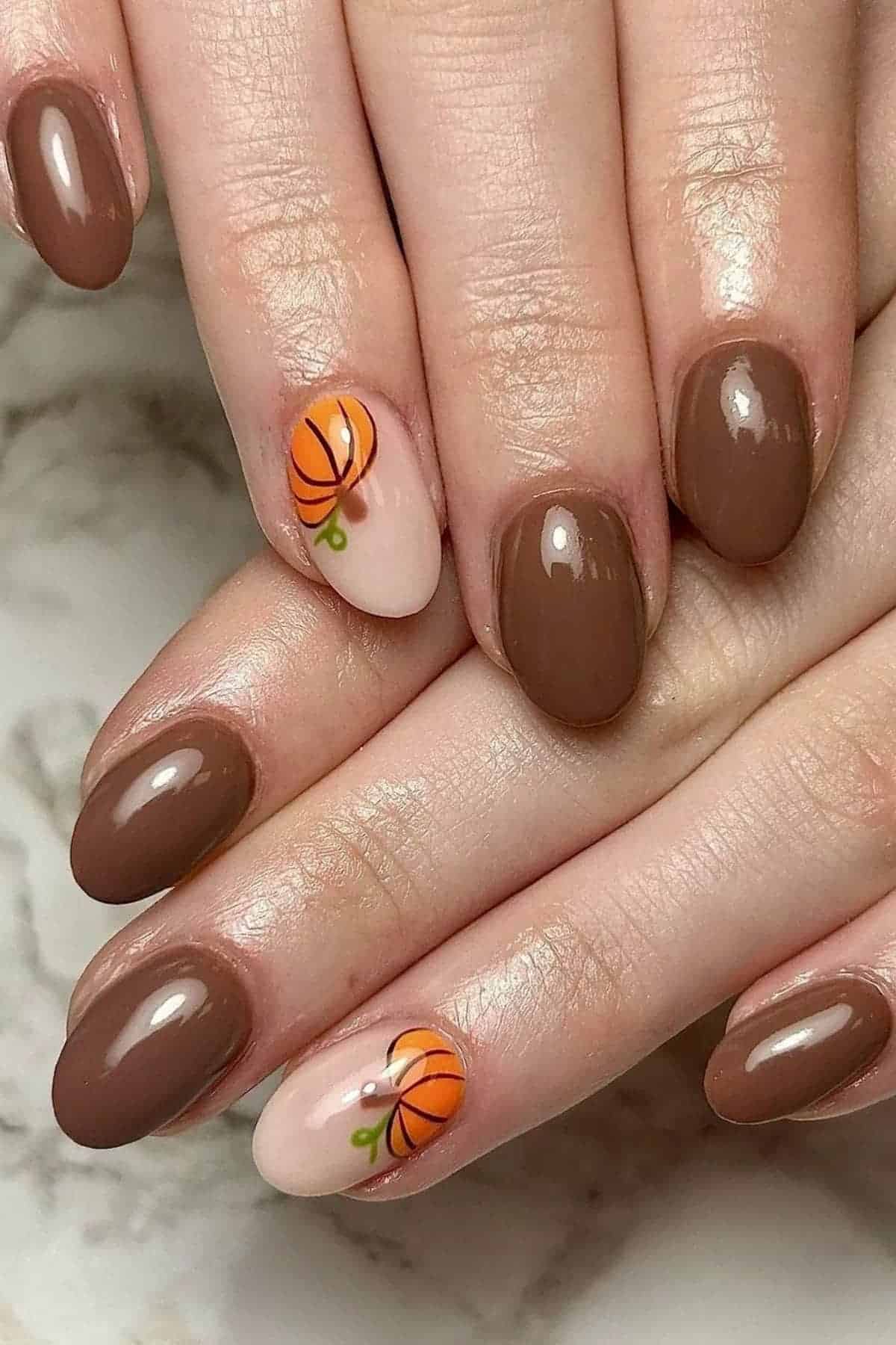 Hands with oval-shaped nails painted brown, with two accent nails featuring an orange pumpkin design on a nude base.