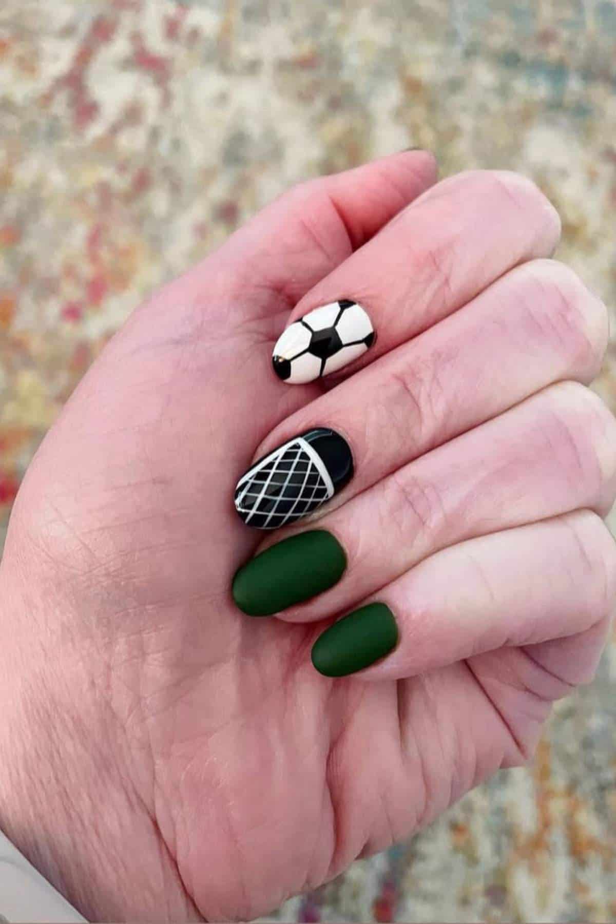 A hand with a manicure featuring two green matte nails, one nail painted like a soccer ball, one with a net pattern, and one glossy black nail.