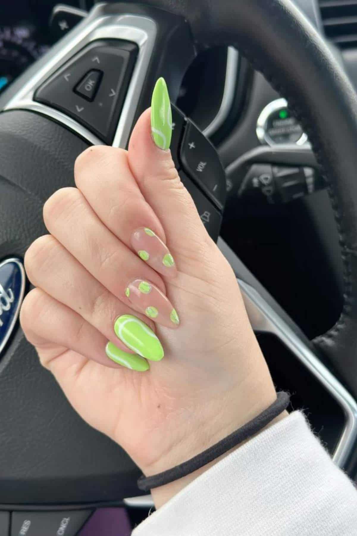 A hand with long, bright green and clear swirl-patterned nails is shown against a car steering wheel with a visible Ford logo.