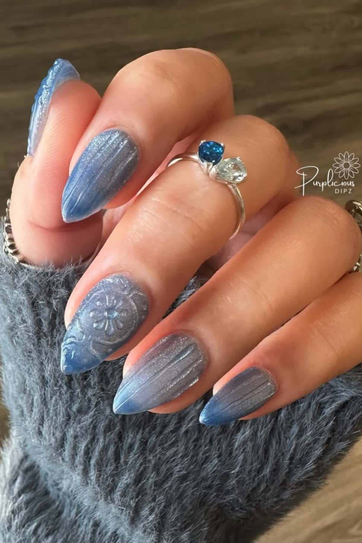 A hand with long, almond-shaped fingernails painted in shimmery blue with a floral accent design, wearing a silver ring with blue and clear stones.
