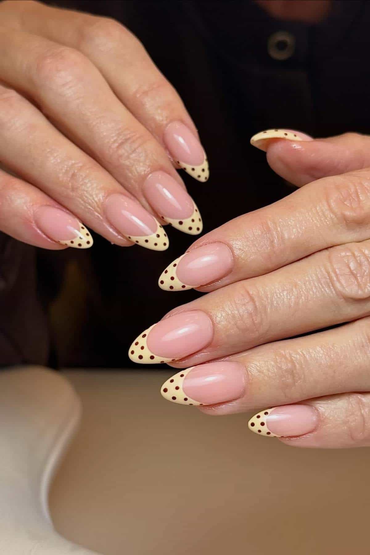 Hands with almond-shaped nails featuring a nude base and cream-colored French tips with brown polka dots.