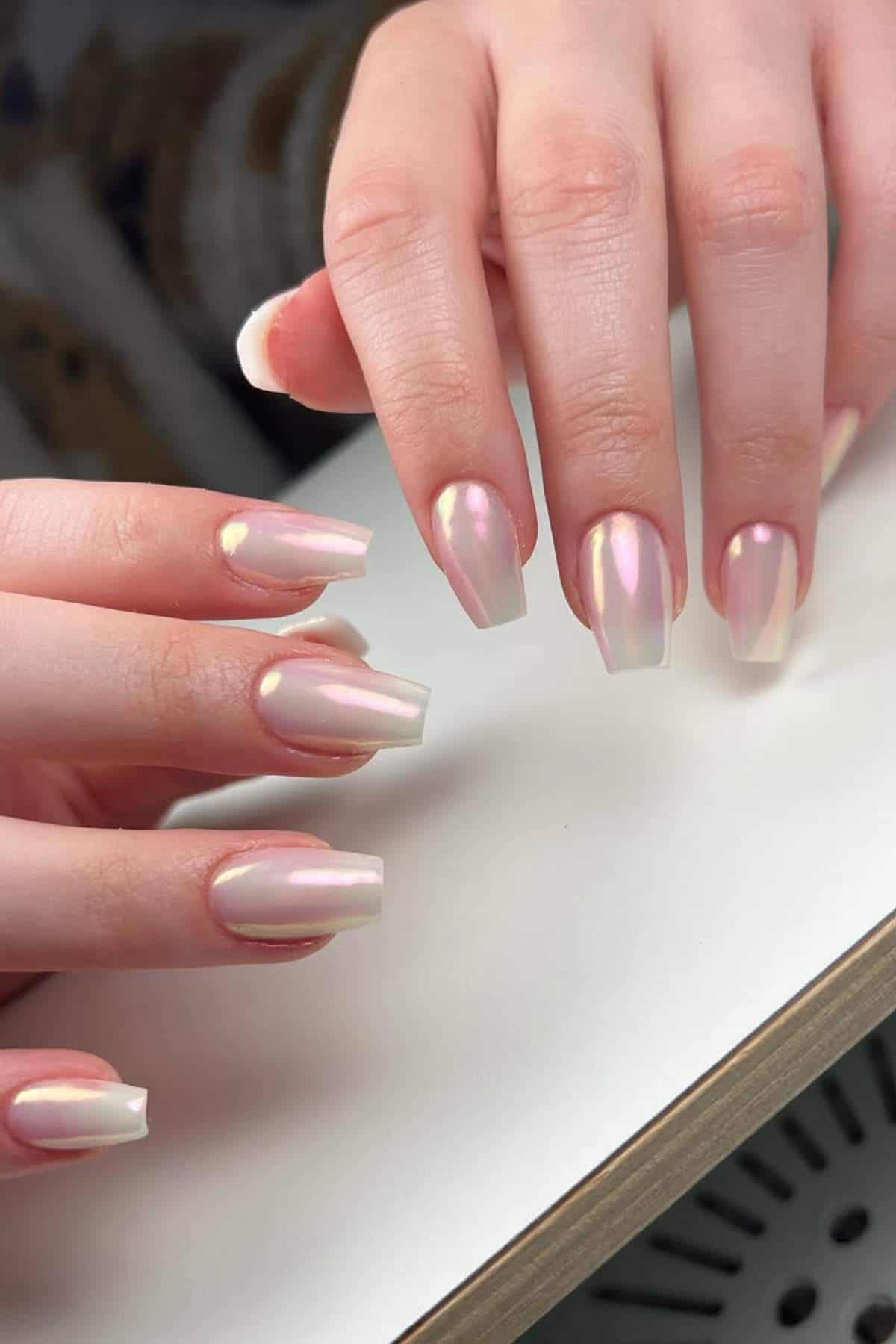 Close-up of hands with medium-length, square-shaped nails painted in a glossy, iridescent pale pink color.