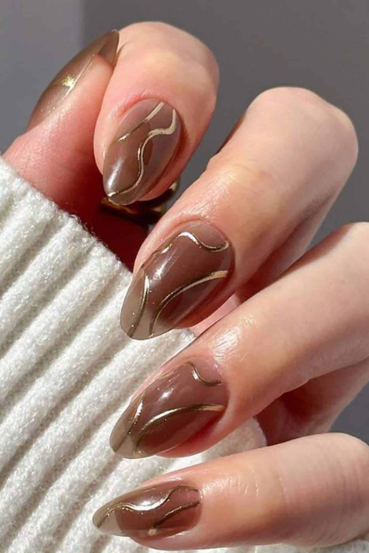 A hand with long, almond-shaped nails painted in glossy brown with gold abstract line designs, shown against a white ribbed sleeve.