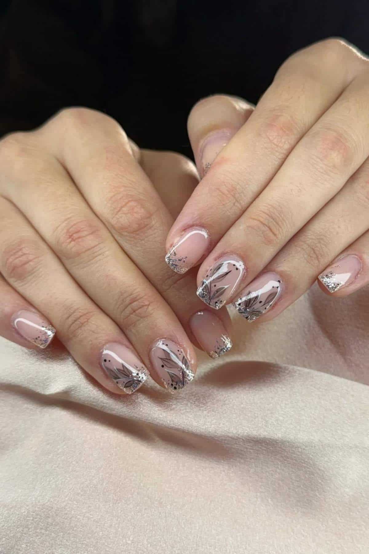Hands with short, almond-shaped nails featuring a pale pink base, silver glitter tips, and black and silver abstract line designs on each nail.