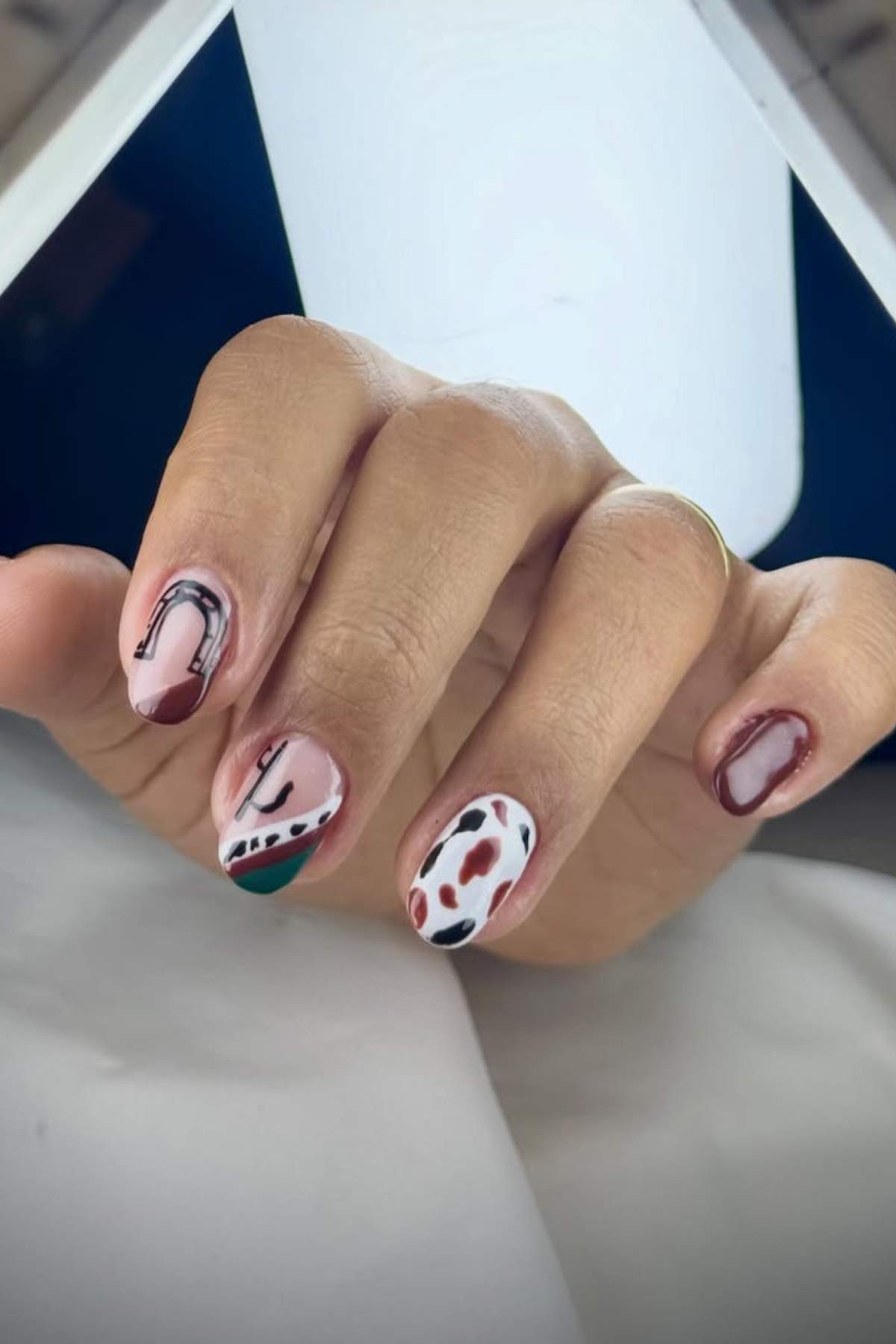 A hand with short nails painted in a mix of abstract designs, including horseshoes, cow print, and brown outlines on a light background.