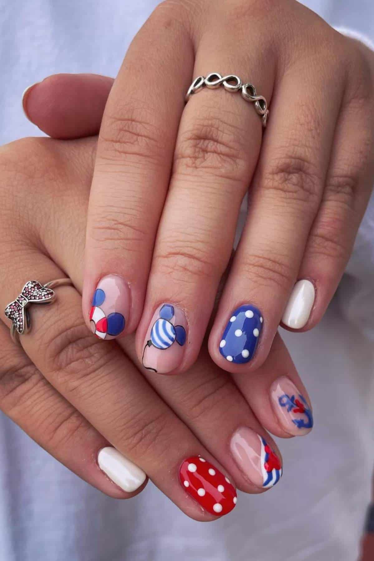 Close-up of hands with manicured nails featuring red, white, and blue designs, including polka dots, stripes, balloons, and abstract patterns. Two silver rings are worn on the fingers.