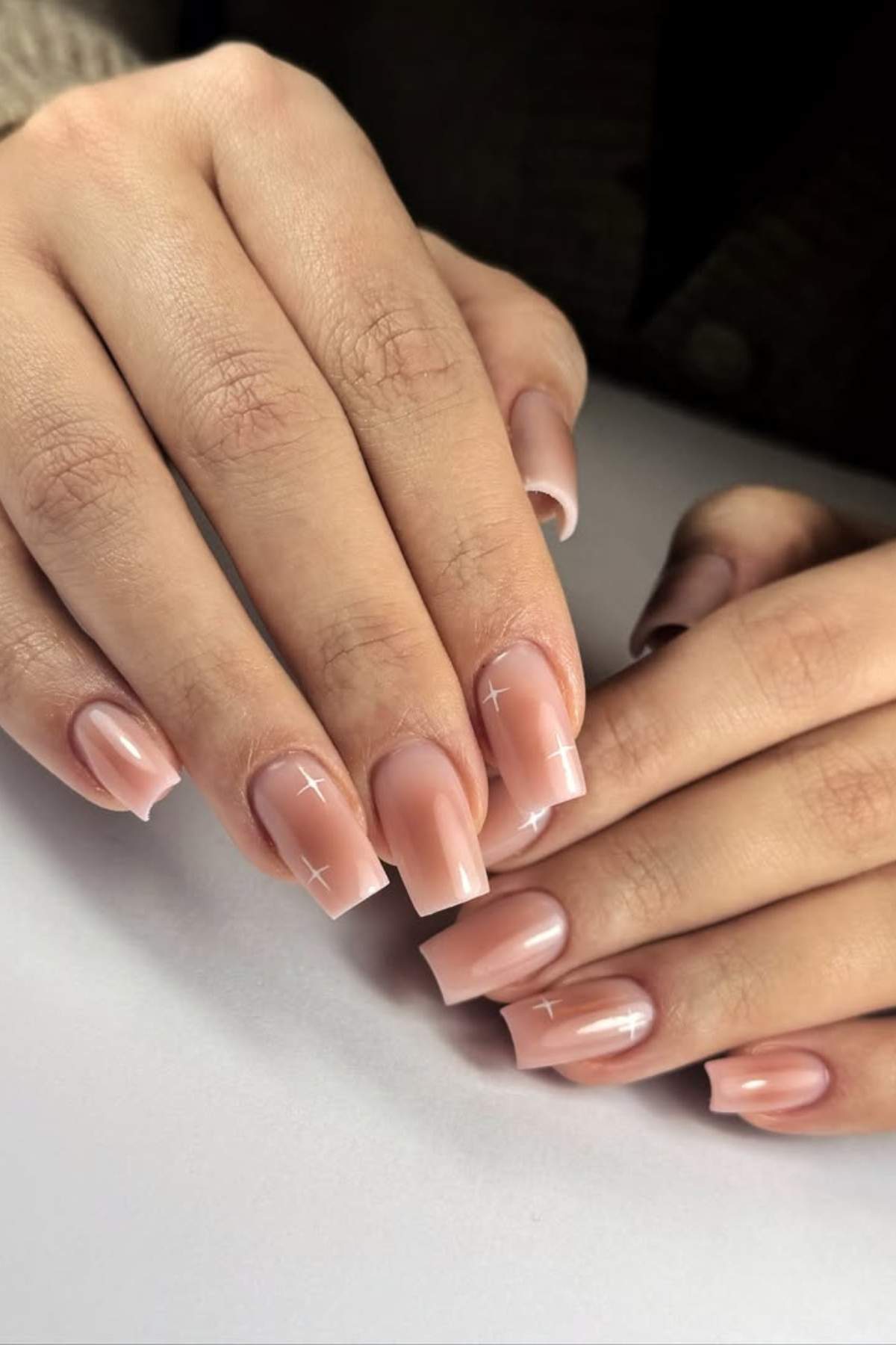 Hands with neatly manicured, square-shaped nails in a soft nude color, featuring subtle white cross accents on a few nails.