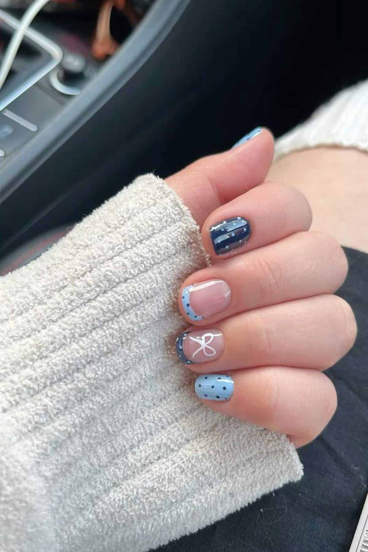 A hand with short, manicured nails painted in a blue and nude color scheme with designs including polka dots, stripes, a bow, and French tips.