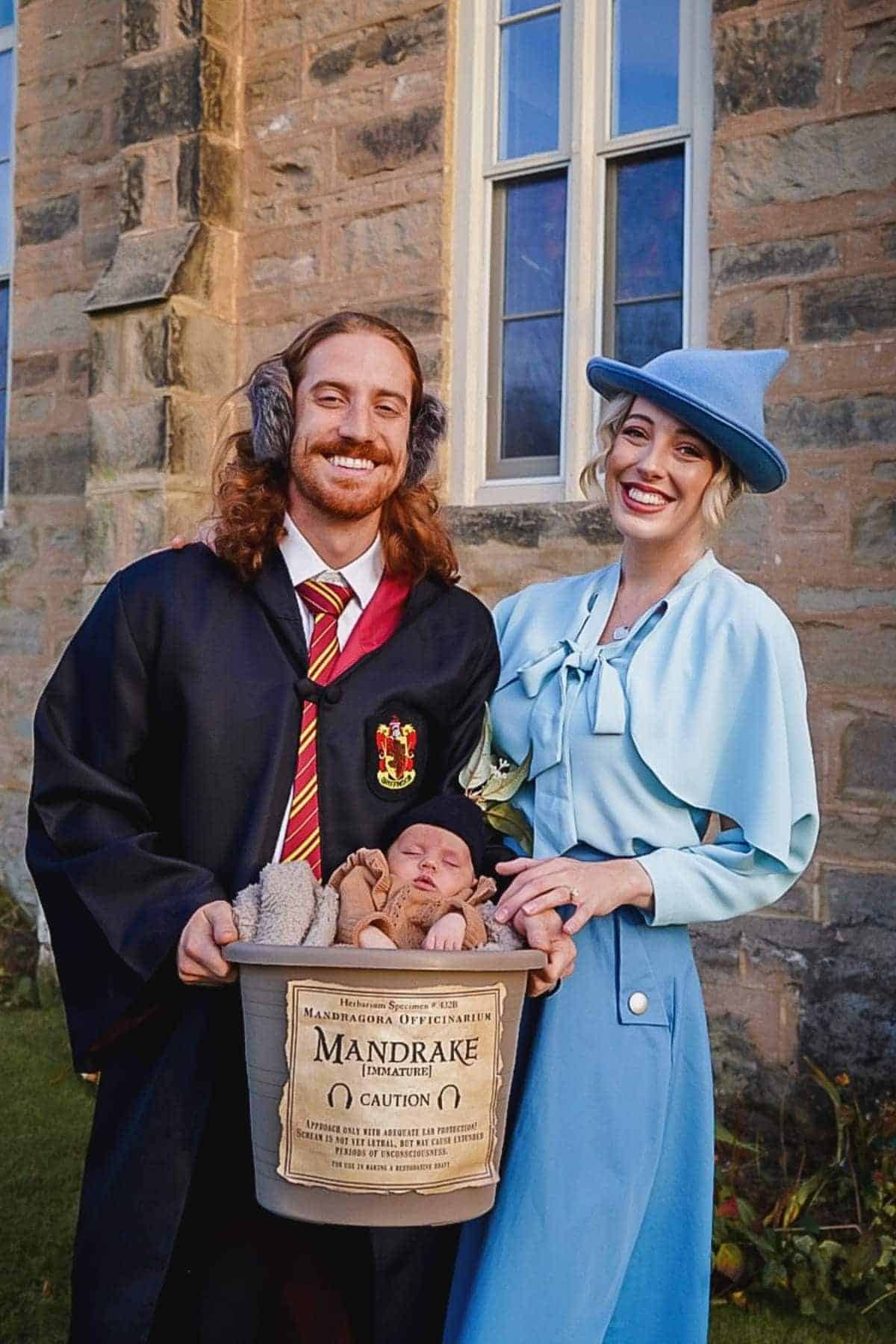 A man in a Gryffindor robe and a woman in a light blue outfit pose outside with a baby in a Mandrake plant prop, referencing Harry Potter.