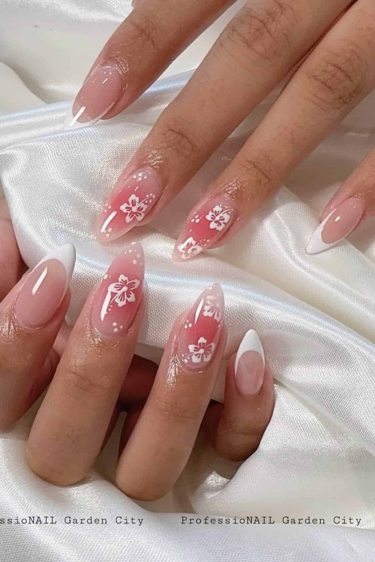 Hands with manicured nails featuring a pink base, white floral designs, and white French tips, displayed on a white satin fabric.