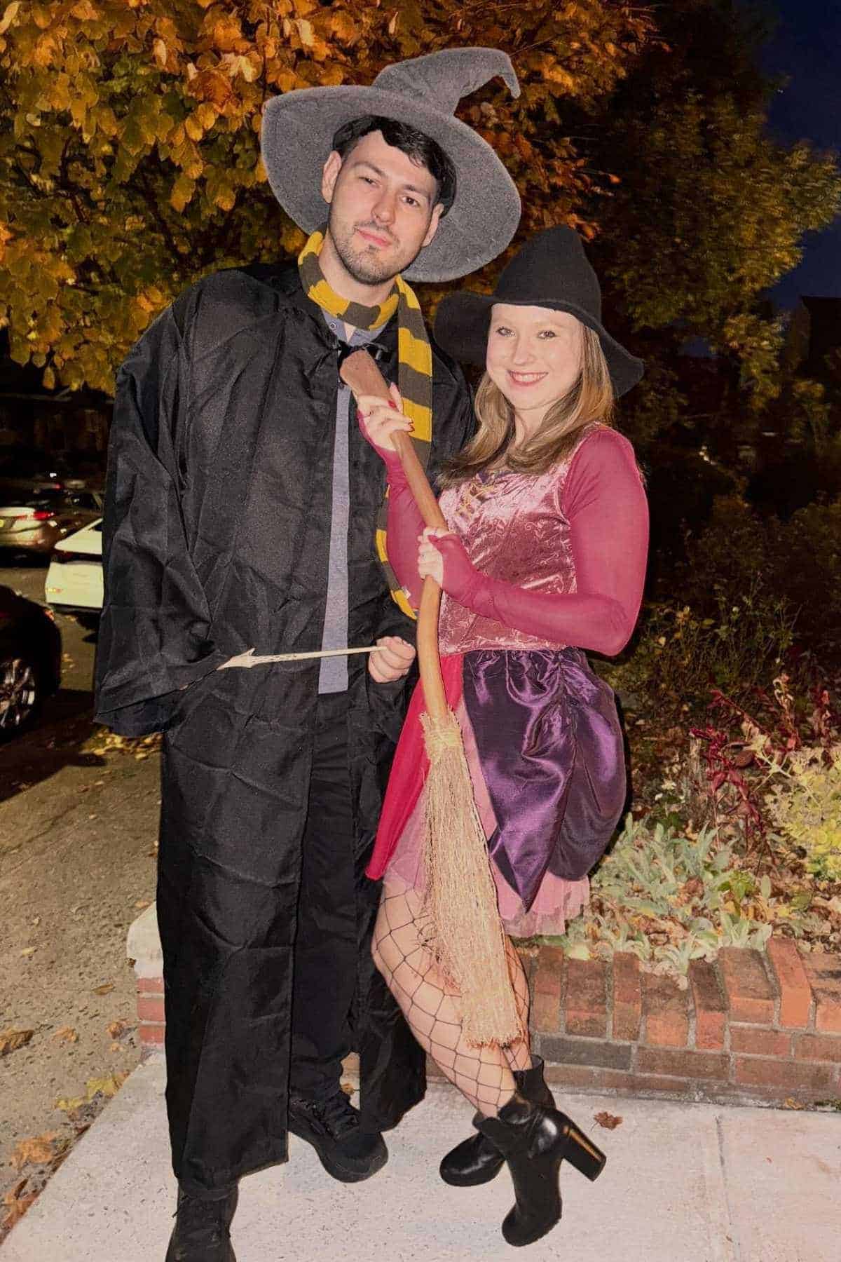 Two people in Halloween costumes pose outdoors at night. One wears a wizard hat and robe, the other wears a witch hat, dress, fishnet tights, and holds a broom. Autumn leaves in background.